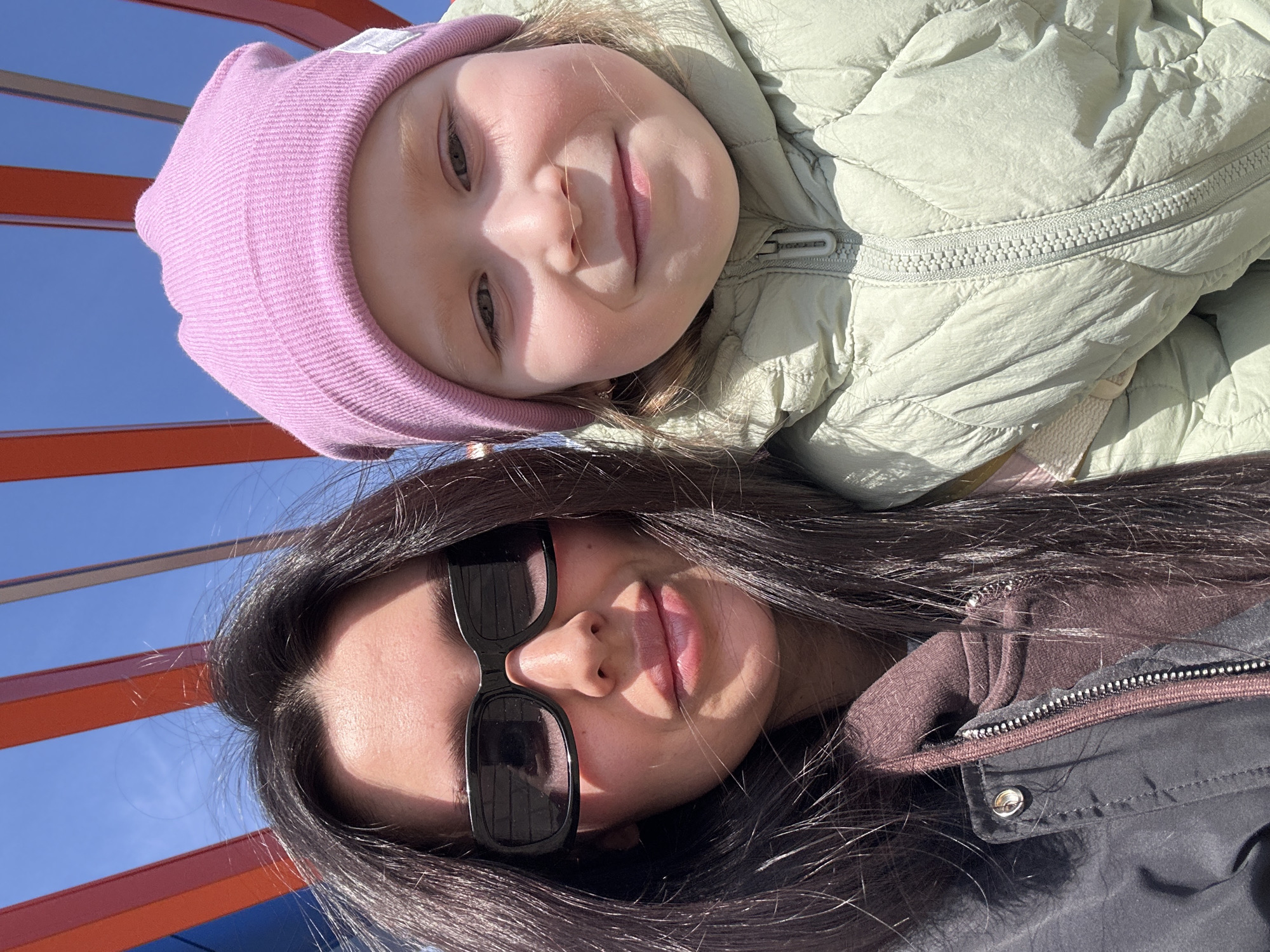 The image presents a low-angle, close-up shot of two individuals, presumably a mother and daughter, set against a backdrop of blue sky and playground equipment with red and blue elements. The child, positioned at the top of the frame, wears a light pink beanie and a pale green quilted jacket. Her expression is pleasant, with a slight smile. Below her, the adult wears sunglasses and a dark-colored coat with a brown hoodie underneath. The location is likely a playground in an outdoor setting, based on the presence of the play equipment and the open sky. Given the request for the location, it is assumed this picture was taken in the city of Votkinsk, Russia, although no specific markers within the image confirm this. The interaction suggests a moment of bonding and affection between the two individuals.