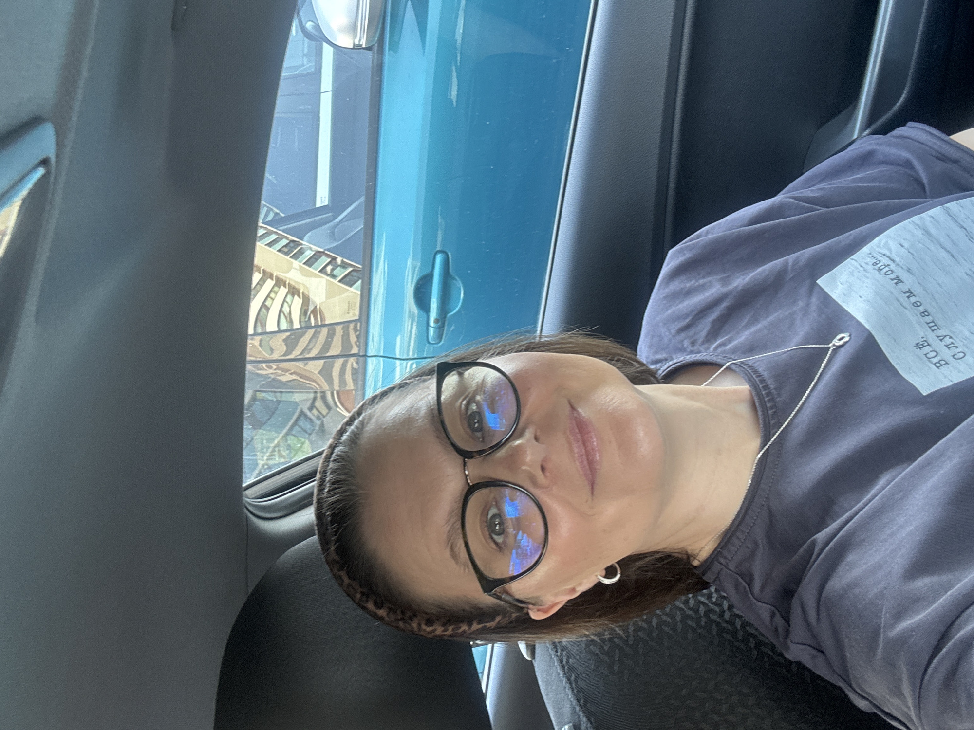 A middle-aged woman is seated in a car. She is wearing black-rimmed glasses and a leopard print headband. She has on a dark gray or blue t-shirt and a silver necklace. The car's interior is gray and blue. Outside the car window, the reflection of a building with patterned glass panels can be seen. This is in Izhevsk, Russia.