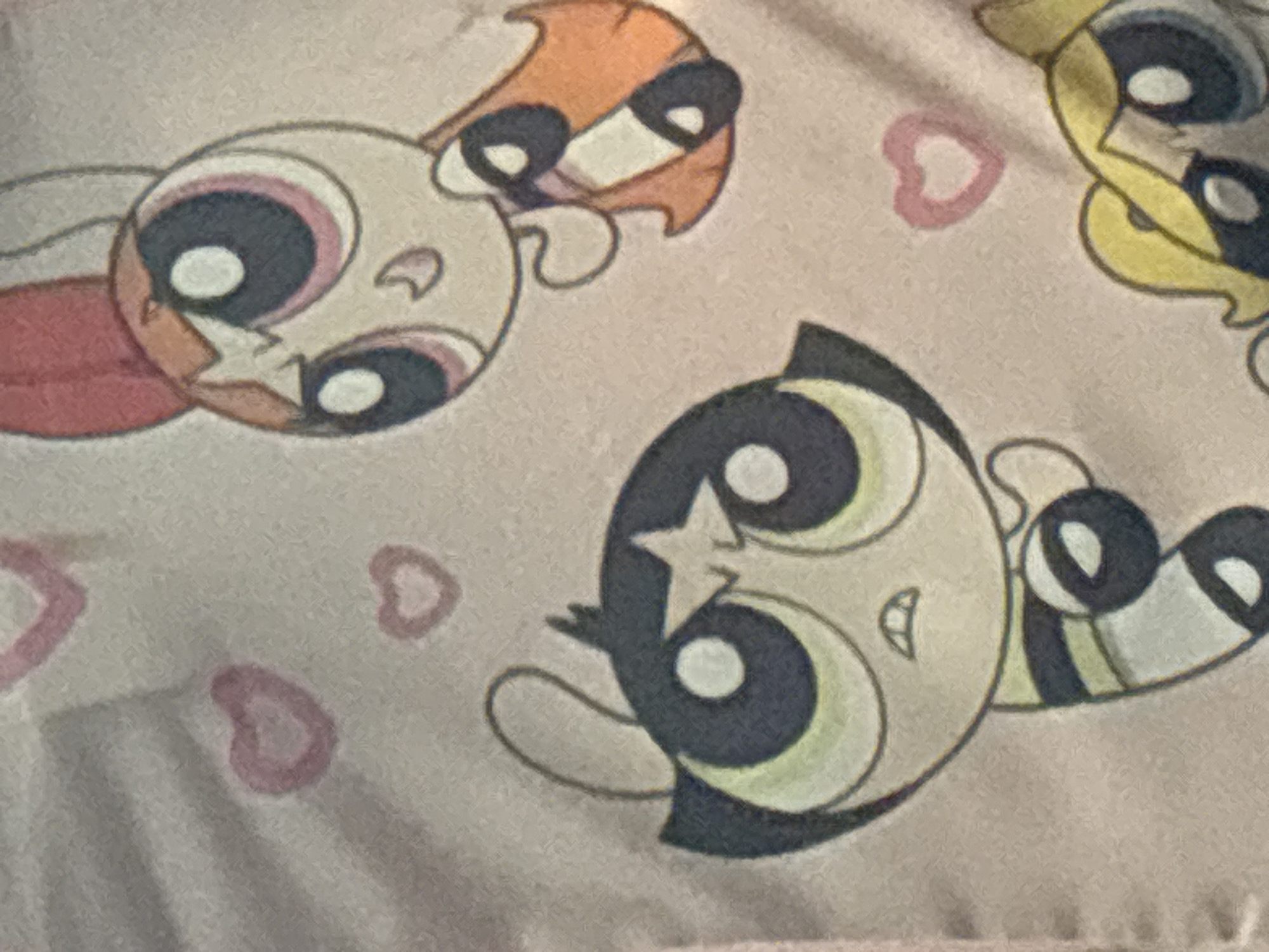 The image is a close-up of a pink textile with a pattern of cartoon characters and hearts. Two of the characters are clearly visible: one with black hair and a star on her forehead, and another with reddish-orange hair and a bow. Both characters have large, round eyes. A third character's blond hair and eye are partially visible at the bottom right. The hearts are small and pink. The textile appears to be fabric, possibly a blanket or clothing. There is no discernible location in Enevo, Bulgaria.