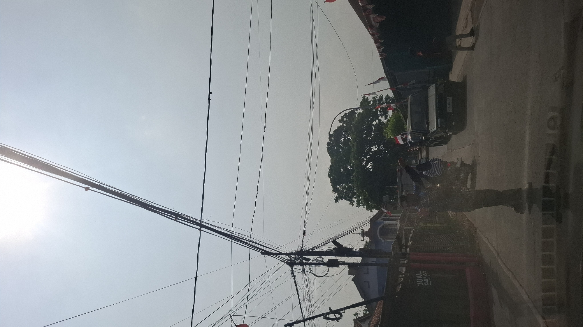 This is a ground-level, outdoor, vertical shot taken on a street in Warungkondang, Indonesia, on a bright, overcast day. The scene is dominated by a dense network of overhead electrical wires crisscrossing the sky. In the foreground, a person in a dark, patterned uniform, possibly a traffic officer, stands in the middle of the road, gesturing with their right arm extended upwards and left arm bent at the elbow. The officer's face is not visible. To their left, a dark-colored building with a red accent wall has a sign that reads "JUAL IKAN MAS" (which translates to "FOR SALE GOLD FISH"). Behind the officer, a motorcycle with a passenger wearing a striped shirt is moving away. Further down the road, a dark SUV is also moving. Along the right side of the road, a building or structure is decorated with numerous small Indonesian flags. A large tree with a full canopy is visible behind the SUV and further down the street, to the left of the flags. The road surface is asphalt. The overall impression is of a street scene with some activity, possibly related to traffic management or a local event.
