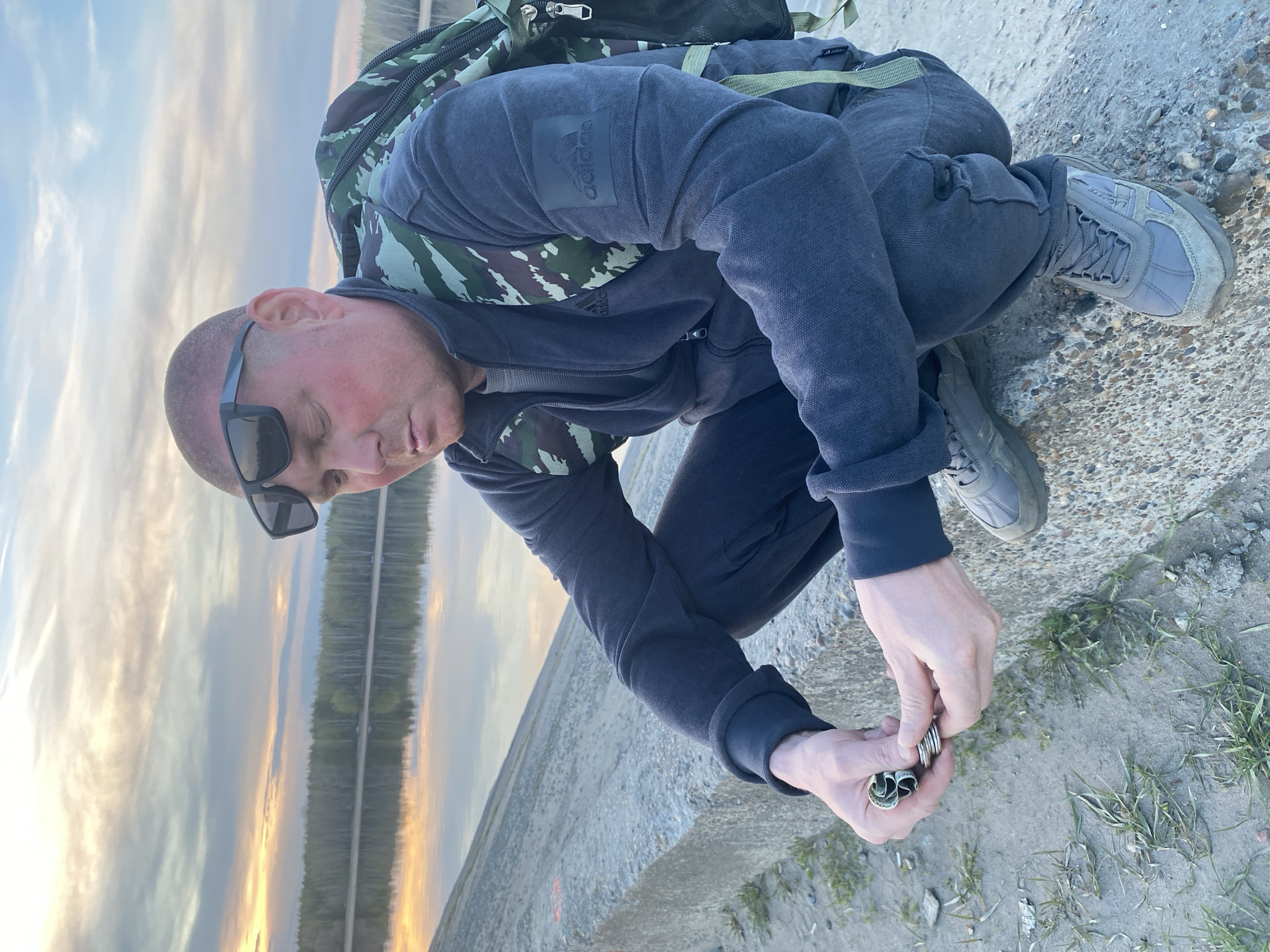 A man with a shaved head and sunglasses is crouching on a concrete structure outdoors, manipulating what appears to be a wad of cash. He is wearing a dark-colored Adidas sweatshirt and sweatpants, with a camouflage backpack behind him. His grey sneakers are visible. The background shows a body of water reflecting a colorful sky, possibly at sunset or sunrise. The location is identified as Noviy, Russia, based on external context.