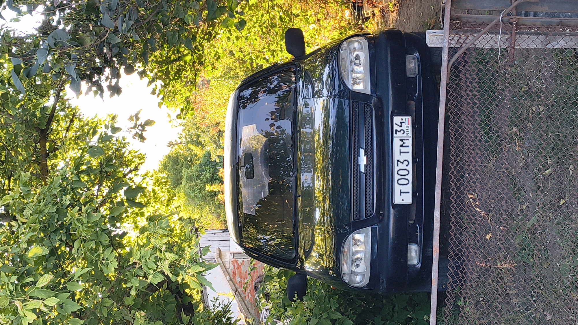 In Zemlyanichnyi, Russia, a dark green Chevrolet Niva is parked on a dirt surface partially obscured by a chain-link fence in the foreground. The car is surrounded by lush green foliage and trees, with sunlight filtering through the leaves, creating a dappled effect. The car's license plate reads "T003TM 34 RUS". Behind the car, a brick structure and more trees are visible, suggesting a rural or semi-rural setting. No people are present in the image.