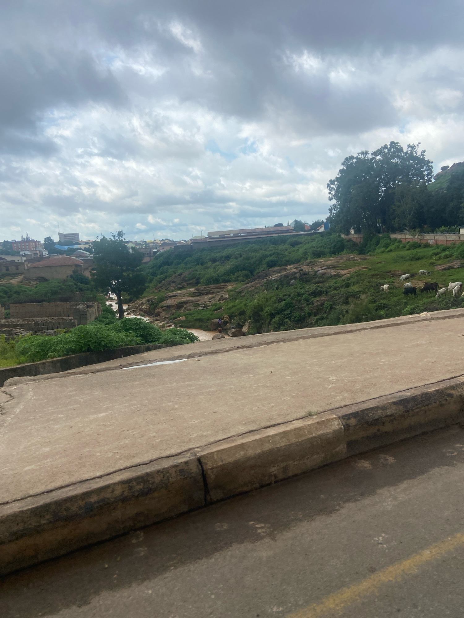 An elevated, slightly tilted, wide shot captures a cloudy day in Jos, Nigeria, with a focus on a rural landscape merging into a town. In the foreground, a weathered concrete road with a raised curb runs diagonally from lower left to upper right. The curb is stained and shows signs of age, with a thin dark line marking its edge. Beyond the road, a steep, rocky incline covered in lush green vegetation slopes downwards.
A small, muddy river winds its way through the terrain. On the far bank, a cluster of cattle, including white and brown individuals, graze on the green slopes. Further up the hill, part of the town of Jos is visible, with buildings ranging from traditional brick structures to more modern, multi-story edifices. A few trees dot the landscape, including a prominent dark green tree in the mid-ground. The sky is filled with heavy, grey clouds, suggesting impending rain or overcast conditions. To the right of the river, a few figures can be seen interacting near the bank, their specific actions are unclear. On the left side of the river, a large pile of bricks is stacked, indicating construction or brick-making activity. The overall impression is of a scene where rural and urban environments coexist, under a dramatic, cloud-filled sky.