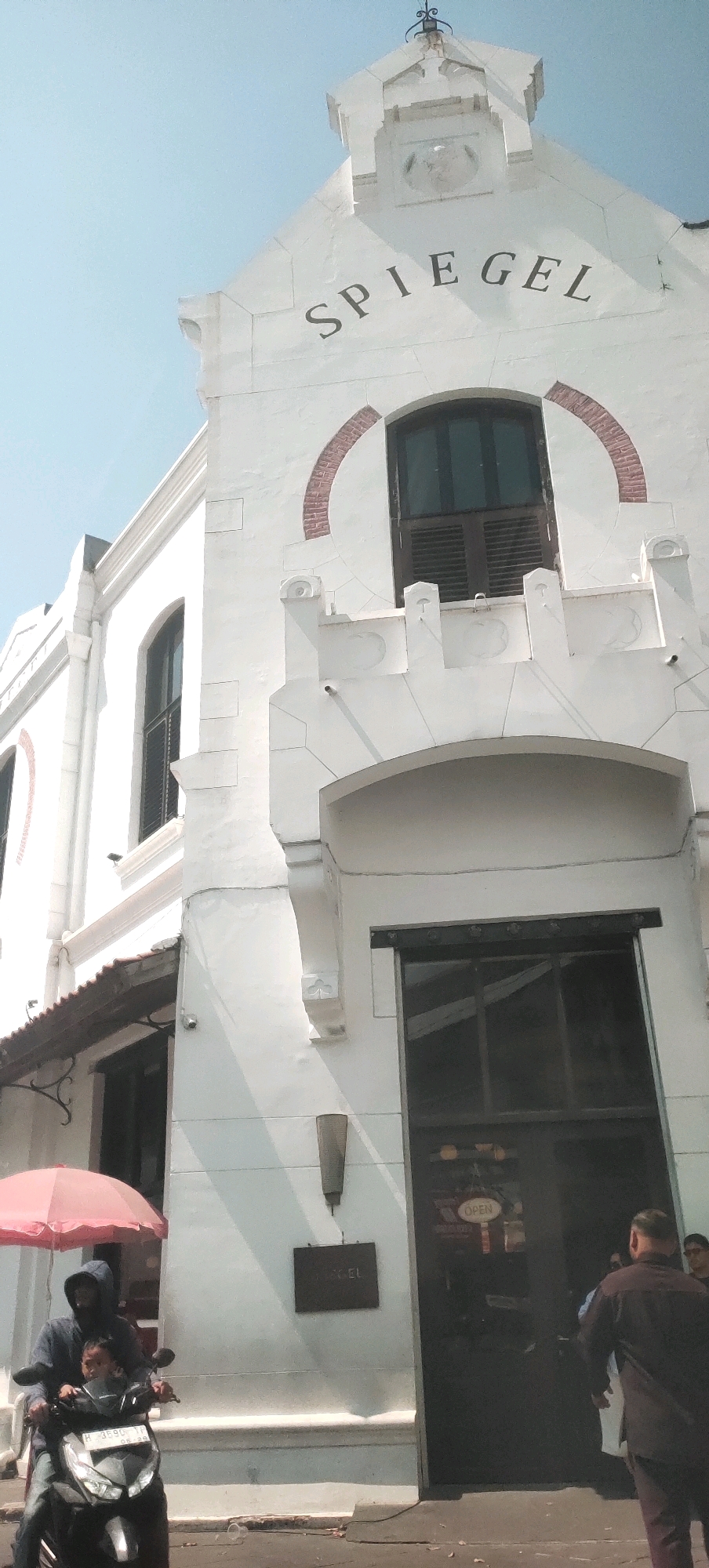 The image depicts the exterior of a white, multi-story building with "SPIEGEL" inscribed in black lettering across its upper facade. The architecture suggests a historical or colonial style, with arched window frames and decorative stone elements. A dark-paned window with wooden shutters is visible in the upper portion of the building. Below this, a darker, arched doorway with large glass panels is present, with a sign reading "OPEN" visible inside.
In the foreground, a person wearing a hooded jacket is riding a scooter, with another individual, possibly a child, seated behind them. A pink umbrella is held over them, providing shade. To the right of the scooter, two individuals are partially visible, one in dark clothing and another in lighter attire. A dark sign is mounted on the wall to the left of the entrance. The overall scene suggests an urban environment with a clear sky. This building is located in the Old City of Semarang, Indonesia, specifically at Spiegel Bar & Bistro.