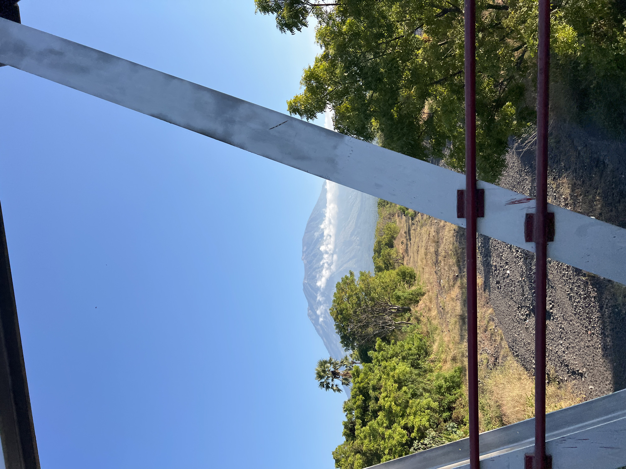 The image captures a scenic view of Mount Agung in Kubu, Indonesia, from what appears to be a bridge or elevated walkway. In the foreground, two red metal railings are visible, attached to white structural supports of the bridge. Beyond the railings, a dirt path with scattered rocks runs through dry grass and low vegetation. A dense line of green trees borders the path on the left, with a solitary palm tree standing tall amidst them. Further in the distance, the majestic Mount Agung is prominently featured, its peak partially shrouded by white clouds. The sky is a clear, vibrant blue, suggesting a bright, sunny day. The overall impression is one of natural beauty and tranquility.