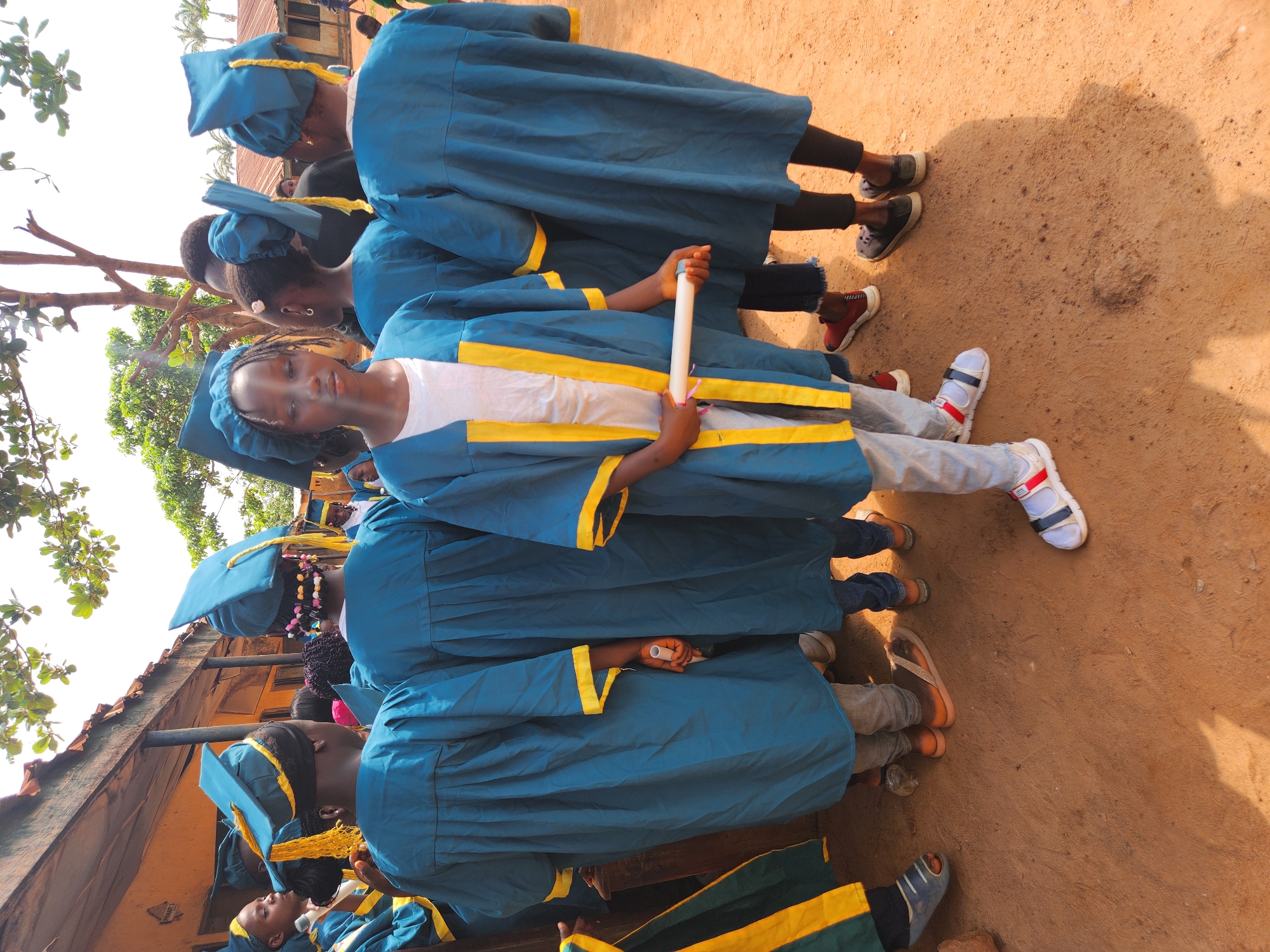 In Sabon Tasha, Nigeria, a group of children in graduation attire stand on a dirt ground in front of a building. The children are wearing blue gowns with yellow trim and matching graduation caps. The central figure, a young girl, holds a rolled-up certificate in her hands. The environment appears to be an outdoor school or community gathering, with a wooden structure partially visible in the background and trees overhead. The overall scene captures a graduation ceremony or a similar celebratory event.