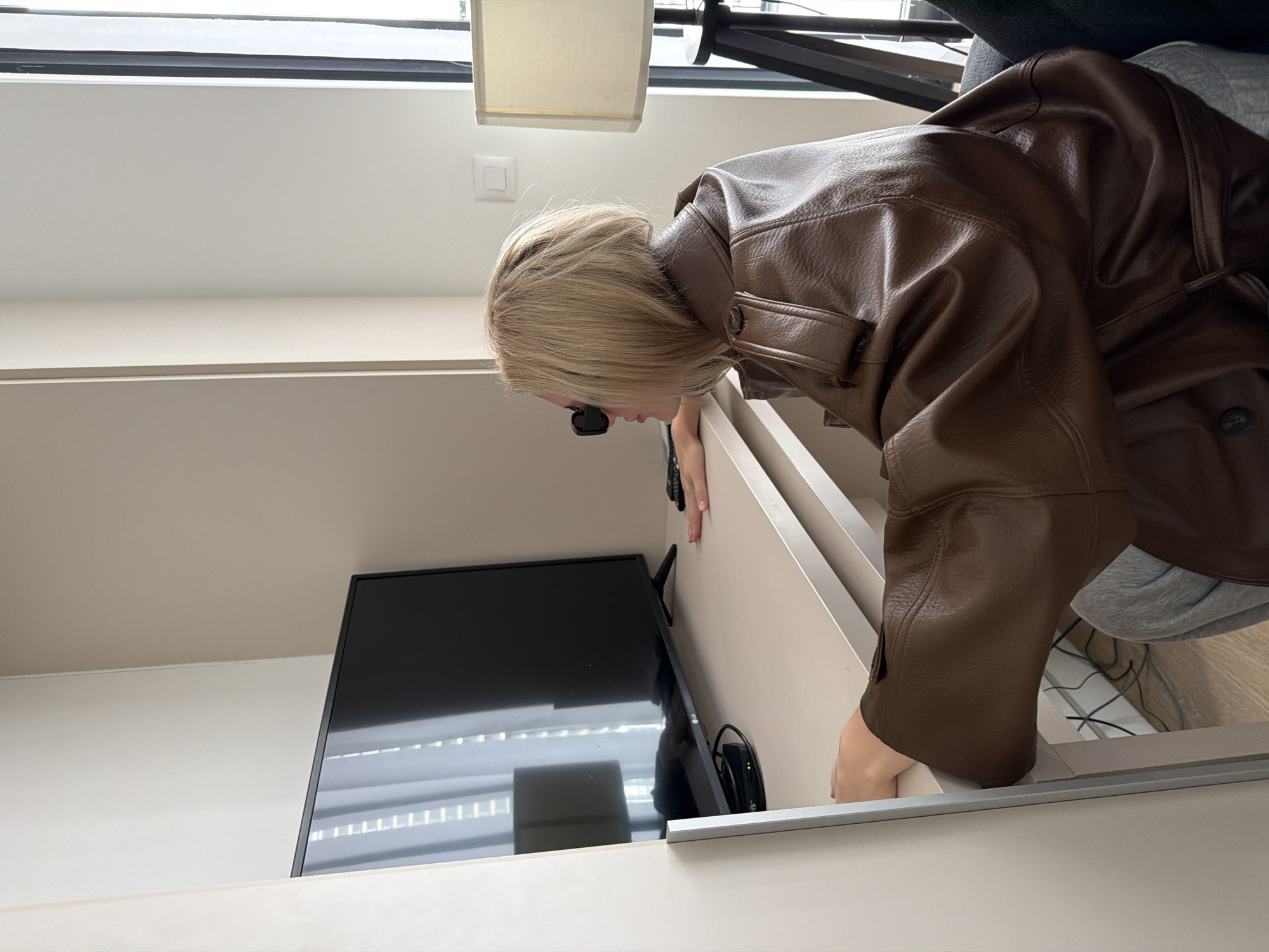 An individual with blonde, bob-length hair and dark sunglasses is seated indoors, leaning forward at a light-colored desk. They are wearing a brown, leather-like trench coat over grey sweatpants. Their hands rest on the desk surface, and their head is turned left, observing a black LG flat-screen television positioned on the desk. Below the television, a small black set-top box with a green indicator light is visible, alongside cables. A black remote control lies on the desk near the person's right hand. To the individual's right, a dark-colored floor lamp with a light-colored, rectangular lampshade stands next to a window, partially showing vertical blinds. The room's walls are light-colored, and the flooring is light wood laminate. An electrical outlet or switch is visible on the wall behind the lamp. The specific location within the city of Belgrade, Serbia, is not discernible from the image.