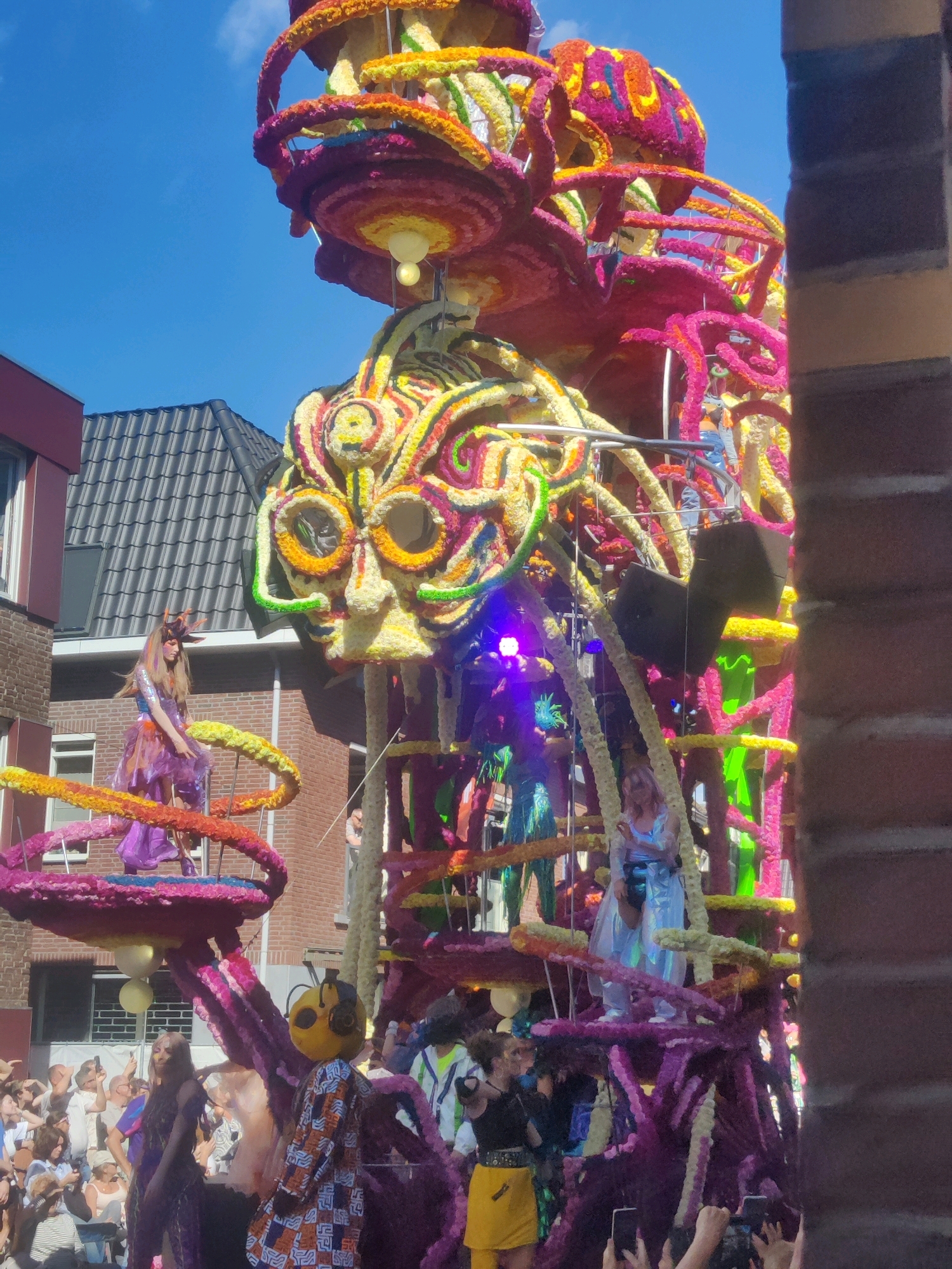 The image captures a parade float in Zundert, Netherlands, during a sunny day. The float is a massive, elaborate structure made of colorful flowers and other materials, depicting a large, intricate, face-like sculpture at the top, with spiraling floral elements extending downwards. Several people are visible on and around the float, some in costumes. In the foreground, a person in a bee-like costume with a patterned outfit is standing. Further back, a woman in a purple outfit with a headpiece is positioned on a spiral platform. Other individuals, some in colorful outfits, are present on the float and in the crowd watching the parade. The background shows buildings with dark roofs and brick facades, and a clear blue sky. The scene suggests a festive event, likely the annual Bloemencorso in Zundert, which is famous for its large floral floats.