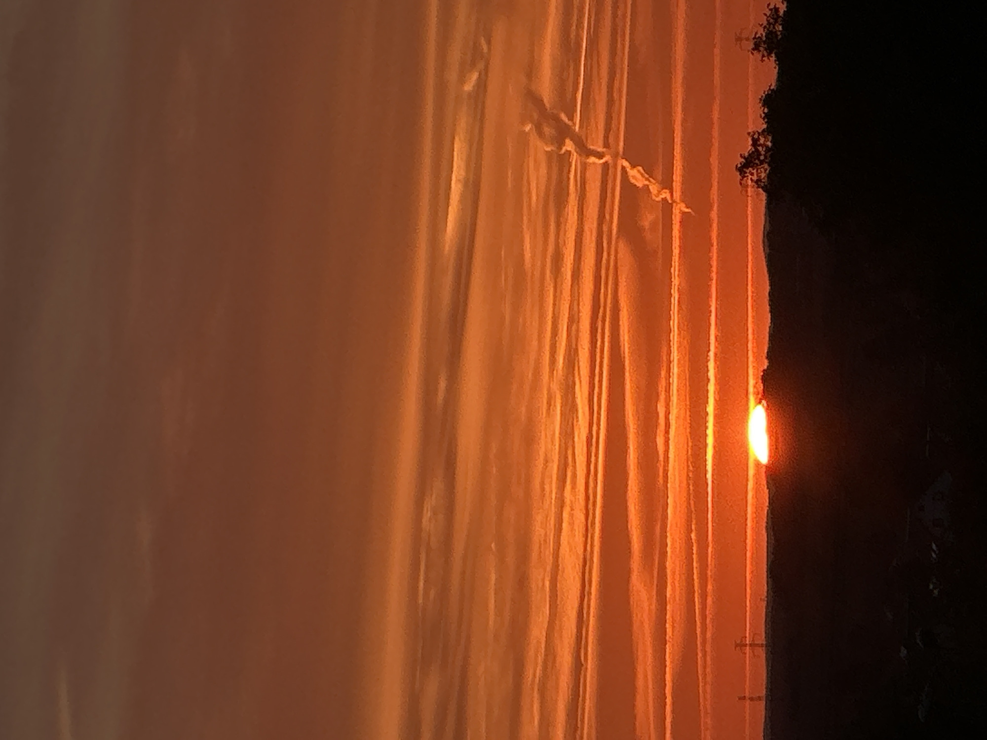 The image captures a dramatic sunset over Świdnica, Poland. The sky is a vibrant canvas of oranges and reds, with distinct horizontal cloud layers streaking across the frame. A single, bright sun is setting on the horizon, partially obscured by a dark, silhouetted ridge that marks the city's landscape. In the distance, a power line tower is visible against the colorful sky, adding a touch of industrial structure to the natural beauty. Some trees are also silhouetted against the horizon on the right side of the image. The overall impression is one of a tranquil, yet awe-inspiring natural spectacle, viewed from a slightly elevated vantage point within the city.