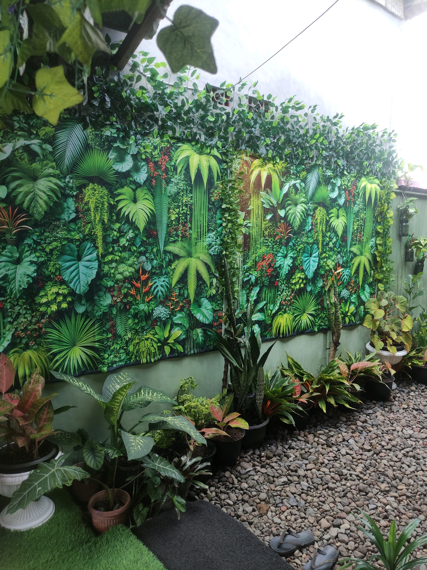 A green wall mural depicting a dense jungle scene with various tropical plants is mounted on a light green wall. The mural features large-leafed plants, ferns, hanging vines, and red flowers. In front of the mural, several potted plants are arranged. These include snake plants, broad-leafed plants with variegated patterns, and plants with pink and green foliage. The ground in front of the mural is covered with gray pebbles, with a dark gray mat on the left and artificial grass on the far left. A pair of gray flip-flops rests on the pebbles. The scene appears to be an outdoor space, possibly a patio or garden area, in Setail, Indonesia. The lighting suggests it is daytime, with sunlight illuminating parts of the scene.