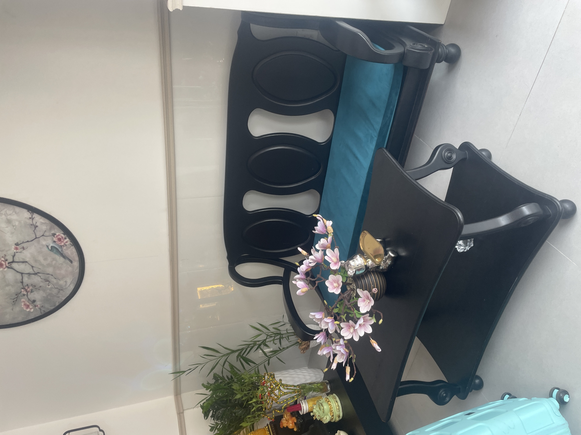 In a bright, modern room, possibly in Thuận An, Vietnam, a sleek black wooden sofa with an ornate back design is the focal point. The sofa is adorned with a vibrant turquoise cushion, offering a stark contrast to its dark frame. In front of the sofa sits a matching black coffee table with a two-tiered design. The lower shelf of the coffee table displays a few clear acrylic or glass decorative items. The top surface of the coffee table is graced by a floral arrangement in a dark, patterned vase. The flowers are a delicate pink, resembling magnolias. Beside the vase, a small, polished gold-colored decorative item rests, possibly a tray or a sculpture.
To the left of the coffee table, a collection of green plants in a white vase adds a touch of nature. Behind the plants, a tall, slim bamboo-like plant stands in a pot, further enhancing the room's natural elements. A hint of other decorative items, including a golden figurine, can be seen on a dark surface behind the plants.
The room's walls are a clean, neutral white, with a decorative circular art piece featuring a branch with pink flowers and a bird hanging on the upper wall. A subtle decorative trim runs along the junction of the wall and the tiled surface. The floor is tiled in a light, neutral color. To the far left, a portion of a bright turquoise suitcase with wheels is visible, suggesting the room might be a waiting area or a part of a travel-related establishment. The overall impression is one of a clean, well-decorated, and possibly commercial or residential space.