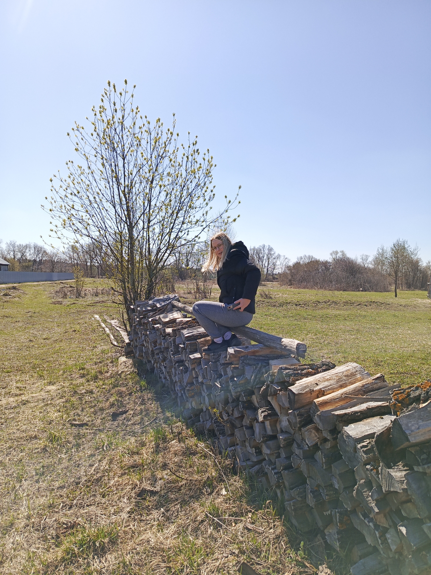 A woman is sitting on a stack of firewood in a grassy field. The woman has blonde hair and is wearing glasses, a black jacket, and gray pants. A tree is behind her to the left. The sky is blue. The location appears to be rural with trees in the distance.