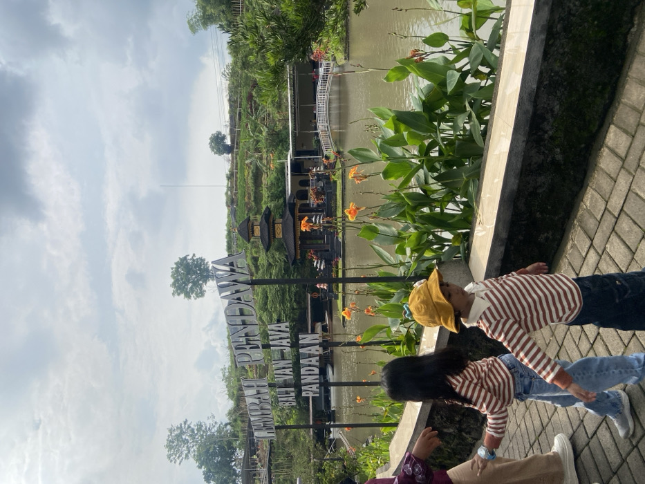 Two children lie on a brick walkway alongside a pond. One wears a yellow hat and a striped shirt, while the other wears a striped shirt and has long dark hair. In the background is a pond with vegetation and a building complex with a sign that reads "LEMBAH PENDAWA BALI VAN JAVA PANDAAN." The sky is overcast.