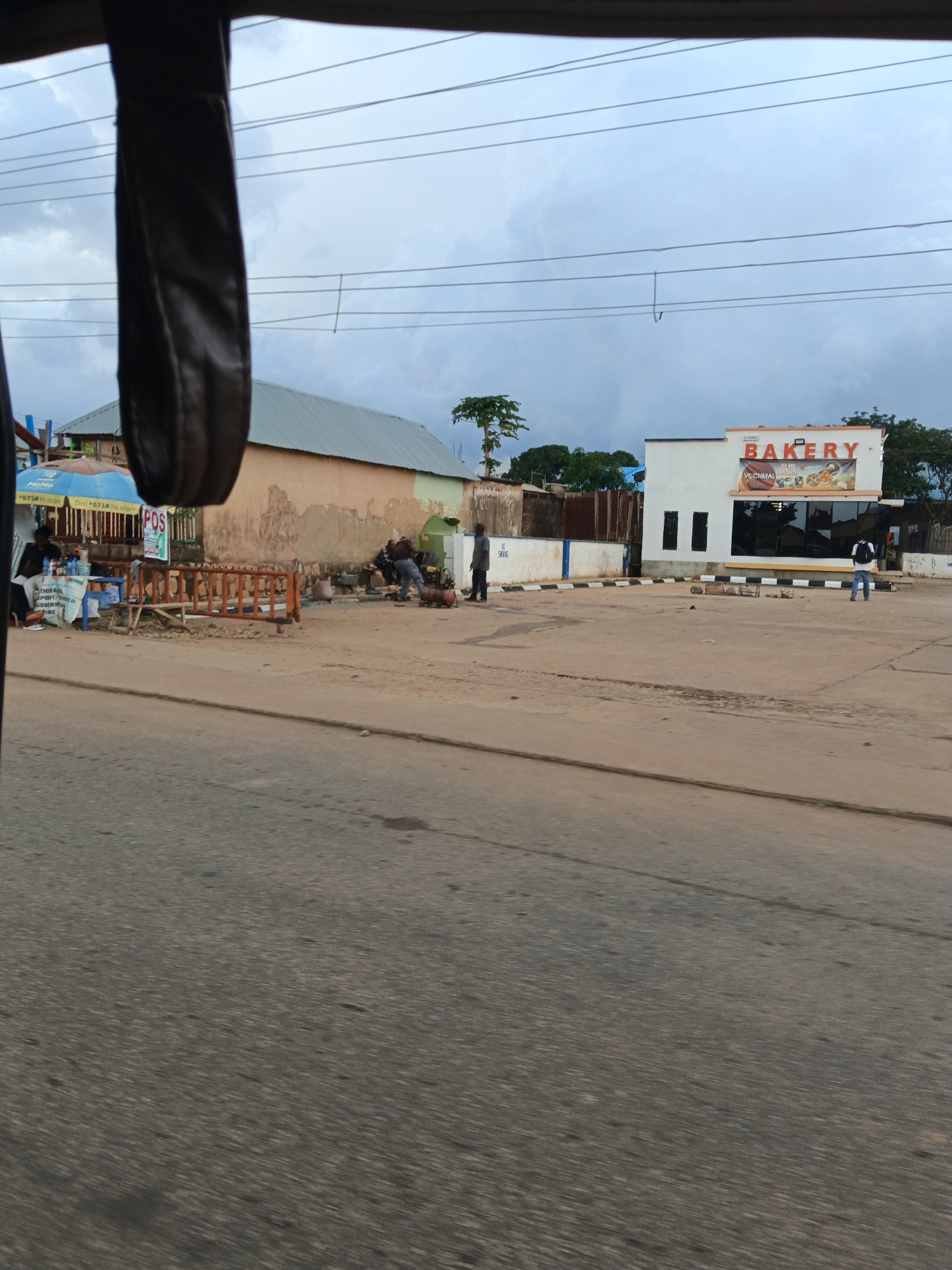 The image captures a street scene in Bukuru, Nigeria, on a cloudy day. In the foreground, a blurred asphalt road dominates the lower portion of the frame, suggesting motion.
To the left, a stall with a blue and yellow umbrella displays various items, including bottles and a visible "POS" sign, indicating a payment terminal. A few individuals are gathered around this stall. Adjacent to it, a low orange barrier with vertical slats separates the stall area from the road.
In the mid-ground, a building with a light-colored, weathered facade is partially visible. Next to it, a modern, white building stands out, featuring large black-framed windows and a prominent "BAKERY" sign in red lettering above a menu board showcasing baked goods. A person wearing a backpack is standing in front of this bakery, facing away from the camera. Another individual in a grey shirt and dark pants is walking towards the right side of the frame, while a person in a dark shirt is standing near the bakery's entrance.
Further back, more buildings and trees are visible under an overcast sky crisscrossed by overhead wires. The overall environment appears to be a commercial area with a mix of older and newer structures.