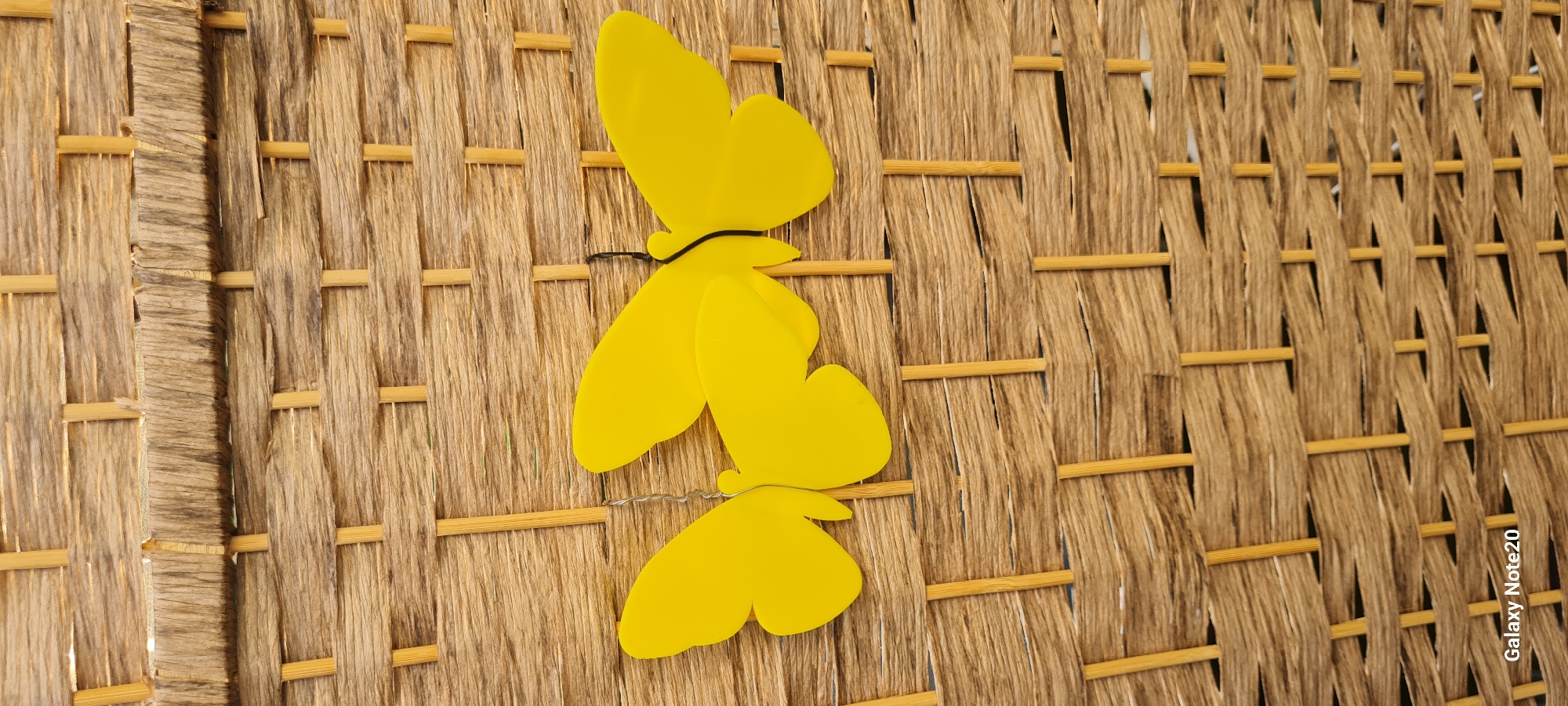 The media file shows a close-up, horizontal shot of a woven texture, likely a basket or mat, made of natural fibers and thin wooden or bamboo rods. Three yellow, stylized butterfly decorations are attached to the woven surface. Two butterflies are stacked vertically, one above the other, and a third butterfly is positioned to the right and slightly above the top one. A thin black cord is visible, used to attach the butterflies. The lighting suggests it is daytime, with shadows indicating a light source from the left. The texture of the weave is prominent, showing variations in color from light brown to darker, more weathered tones, with the horizontal rods providing structure. The setting appears to be outdoors or in a location with natural light. The overall impression is decorative and summery. The text "Galaxy Note20" is visible in white lettering on the bottom right corner, indicating the device used to capture the image. There are no people visible.