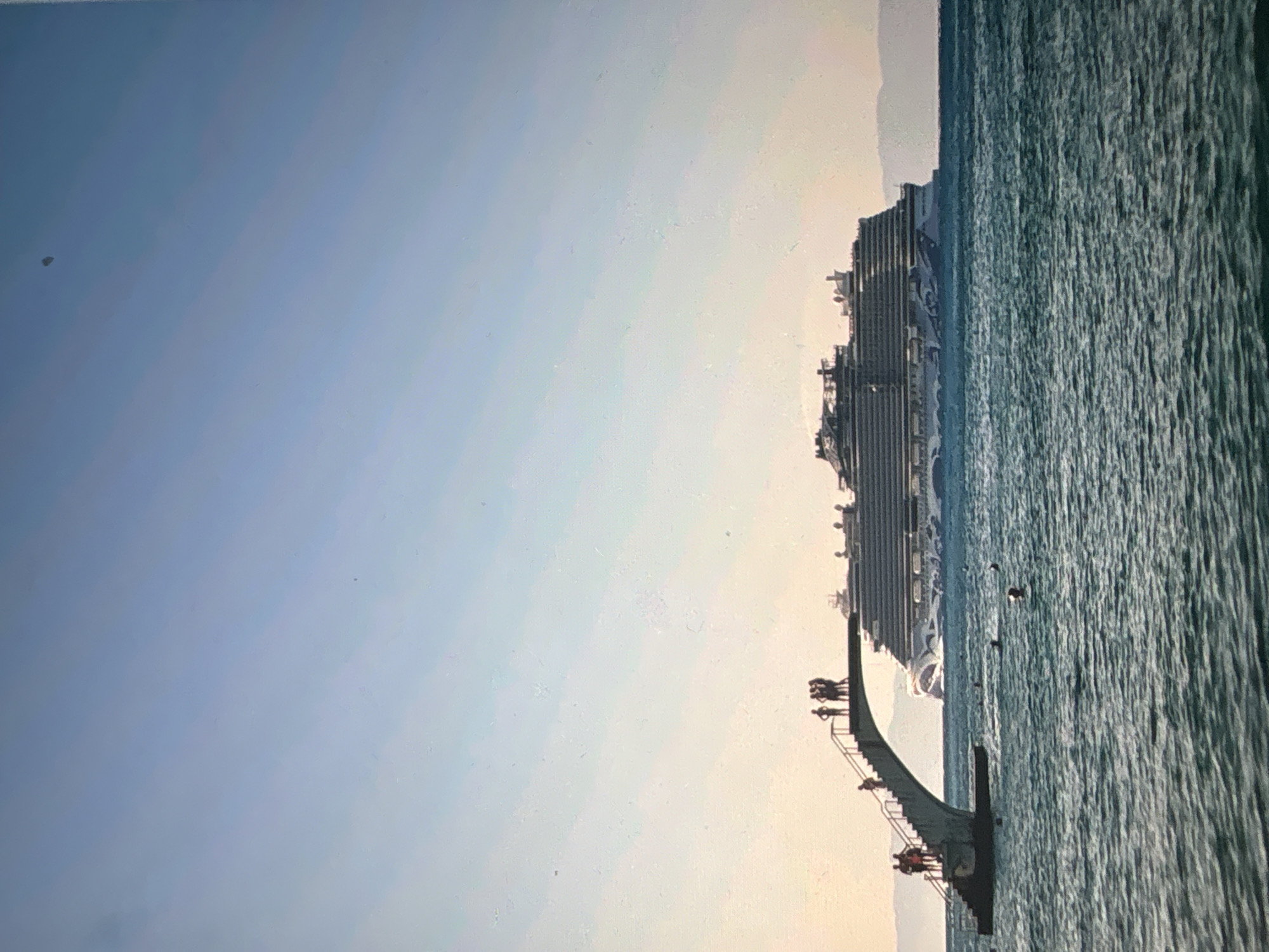 The image displays a wide view of a marine landscape, presented in a portrait orientation. On the left side, a vast, clear blue sky dominates, transitioning to a lighter, hazy pale yellow tone towards the right, indicating a bright horizon. The right side of the frame is filled with a large body of water, likely an ocean or sea, which has a deep teal-blue hue and is covered with numerous small, distinct ripples and waves, suggesting gentle movement.
Stretching horizontally across the mid-section of the image, where the sky meets the water, is a large, multi-decked cruise ship in the distance. Its light-colored hull features visible levels, balconies, and superstructure details. Behind the ship, a faint, distant landmass is discernible along the horizon line.
In the lower-middle foreground, a darker, elongated structure extends horizontally, partially into the water. This structure, which appears to be a pier, jetty, or viewing platform, has railings and multiple visible sections. Several small, silhouetted figures of people are standing or sitting on this structure, seemingly observing the distant ship or the expansive water.
A few small, dark specks, potentially birds, are visible on the water's surface near the foreground structure. Another singular, small, dark, indistinct object is present in the upper-left quadrant of the sky.
The lighting suggests bright daylight, possibly late afternoon or early morning, given the soft glow near the horizon. The weather is clear and calm. The scene evokes a sense of tranquility and observation in a coastal or harbor environment. There is no visible text in the image.