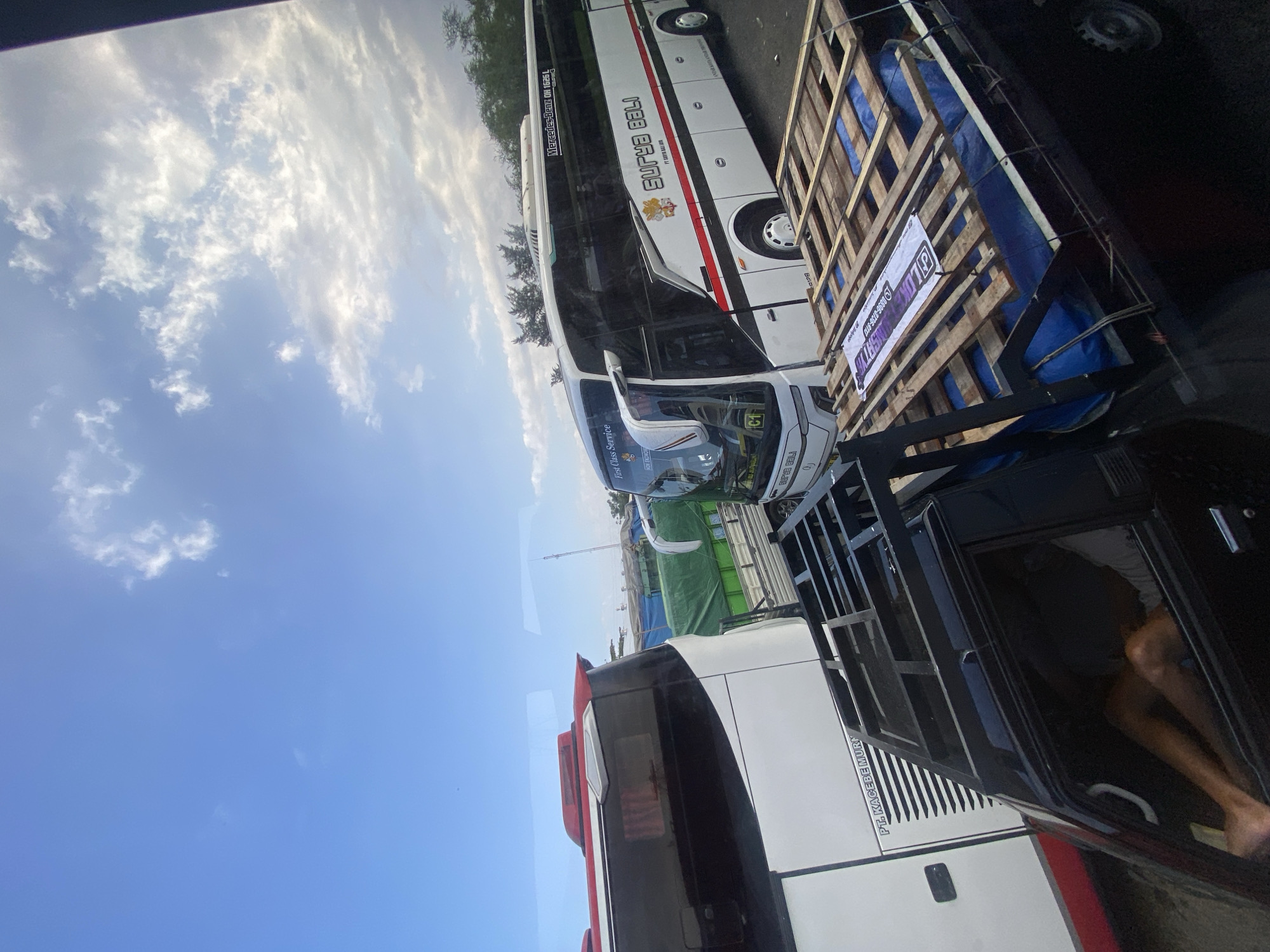 The image depicts a daytime scene, likely in Melaya, Indonesia, characterized by traffic congestion involving several buses and a pickup truck. The sky is partly cloudy, with a mix of blue sky and white clouds, suggesting fair weather.
In the foreground, a black pickup truck is loaded with wooden pallets and covered with a blue tarp. A white banner with text and a phone number is stretched across the pallets. The truck has a visible rack structure on top. Inside the truck's cabin, a person's legs and torso are visible, appearing to be lying down or resting.
Behind the pickup truck, a white and red bus is partially visible on the left. On the right, a white bus with "SURYA BALI" branding is prominently displayed. This bus features "First Class Service" text on its front and has "Mercedes-Benz OH 1625 L" text on its side. Other vehicles, including what appears to be a truck with green tarpaulin cover, are visible further in the background, contributing to the impression of a traffic jam or a transportation hub. Trees and a utility pole are also discernible in the background, indicating an outdoor setting. The overall scene suggests a typical day at a roadside location where transportation is a significant activity.