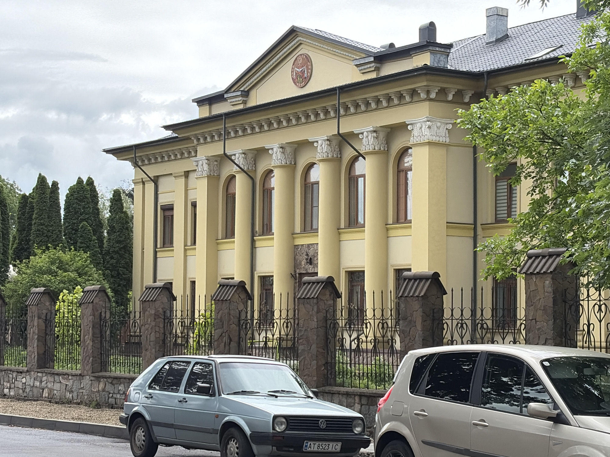 The image depicts the facade of a large, yellow building with classical architectural elements, located in Kolomyia, Ukraine. The building features a colonnade with Corinthian columns, arched windows, and decorative molding. A triangular pediment with a circular emblem is visible at the top center of the facade. The building is partially obscured by a mature tree on the right side of the frame. In the foreground, a light blue Volkswagen Golf Mk2 is parked on the street, facing left, with its license plate AT 8523 IC clearly visible. To the right of the Golf, a silver hatchback car is partially visible. A black metal fence with stone pillars separates the building from the street. The sky is overcast with gray clouds.