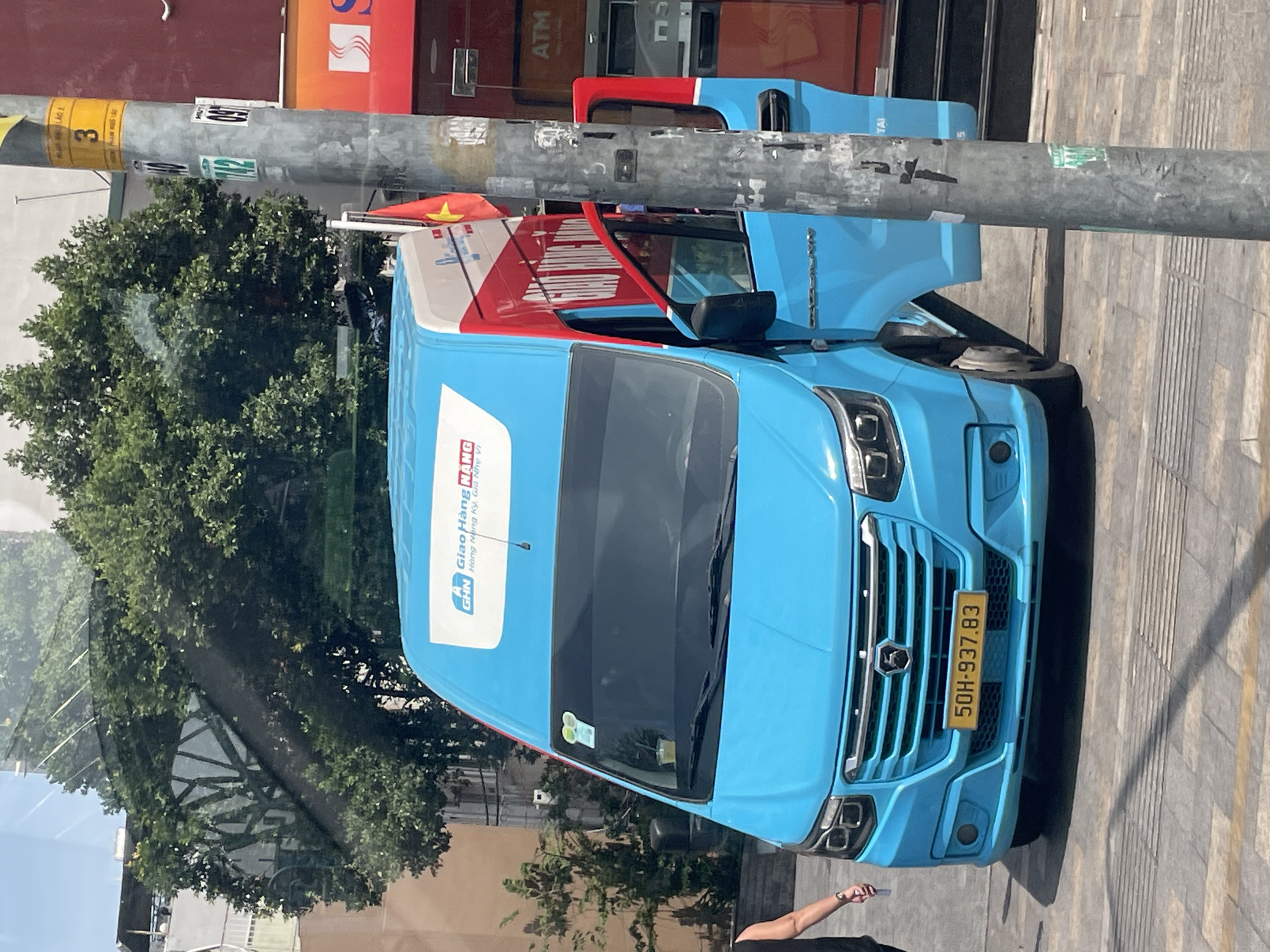 A light blue commercial van with red accents and the "GHN Giao Hàng Nhanh" logo is parked on a city street in Thủ Đức, Vietnam. The van has its driver's side door open, revealing a red interior. A Vietnamese flag is attached to the side of the van. The van's license plate reads "50H-937.83". A person wearing a black tank top and watch is standing to the left of the van. A metal utility pole with stickers and signage is positioned to the right of the van, obscuring part of its side. In the background, trees and buildings are visible, including an ATM sign on a red building. The street is paved with gray and yellow tiles.