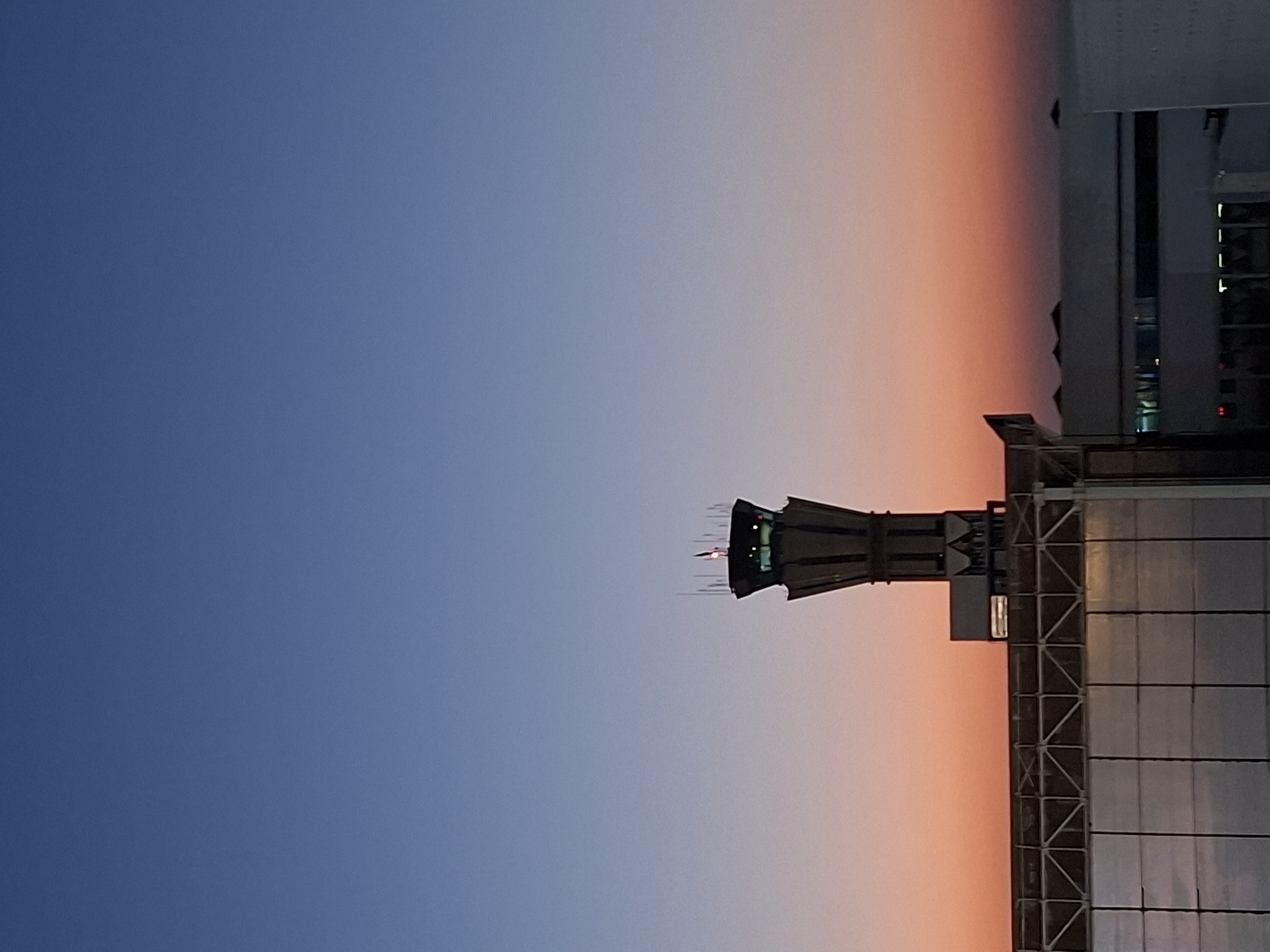 The image captures a view of the Tashkent International Airport control tower at dusk. The sky transitions from a deep blue at the top to a soft orange and pink gradient near the horizon, indicating the end of daylight. The control tower, a prominent vertical structure, stands silhouetted against the colorful sky, its navigational lights faintly visible. In the foreground, the rooftop of airport buildings, characterized by their modern, angular architecture and reflective surfaces, are partially visible. The scene is devoid of people, focusing entirely on the infrastructure of the airport and the atmospheric conditions of the evening sky over Tashkent, Uzbekistan.