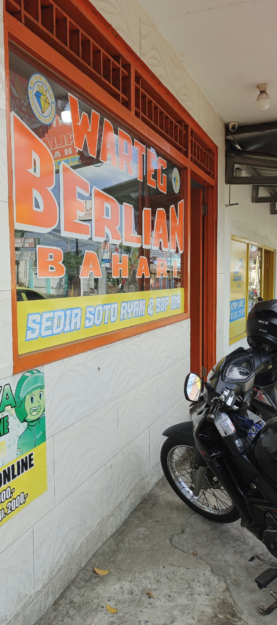 The image captures a street view of a small food establishment, a "Warung," in Yogyakarta, Indonesia. The building's facade is painted white with textured tiles, and the storefront features a prominent orange frame around a large window. The window displays signage for "WARTEG BERLIAN BAHARI" in large, bright orange lettering. Below this, a yellow banner reads "SEDIA SOTO AYAM & SOP IGA," indicating the menu items offered. To the left, a yellow sign with a cartoon character in a green helmet and the "Grab" logo suggests delivery services are available. A black motorcycle is parked in front of the establishment, partly obscuring the lower portion of the storefront. In the background, beyond the window, is a blurred view of a street with other buildings and vehicles. The overall scene depicts a typical local eatery in a bustling urban environment.
