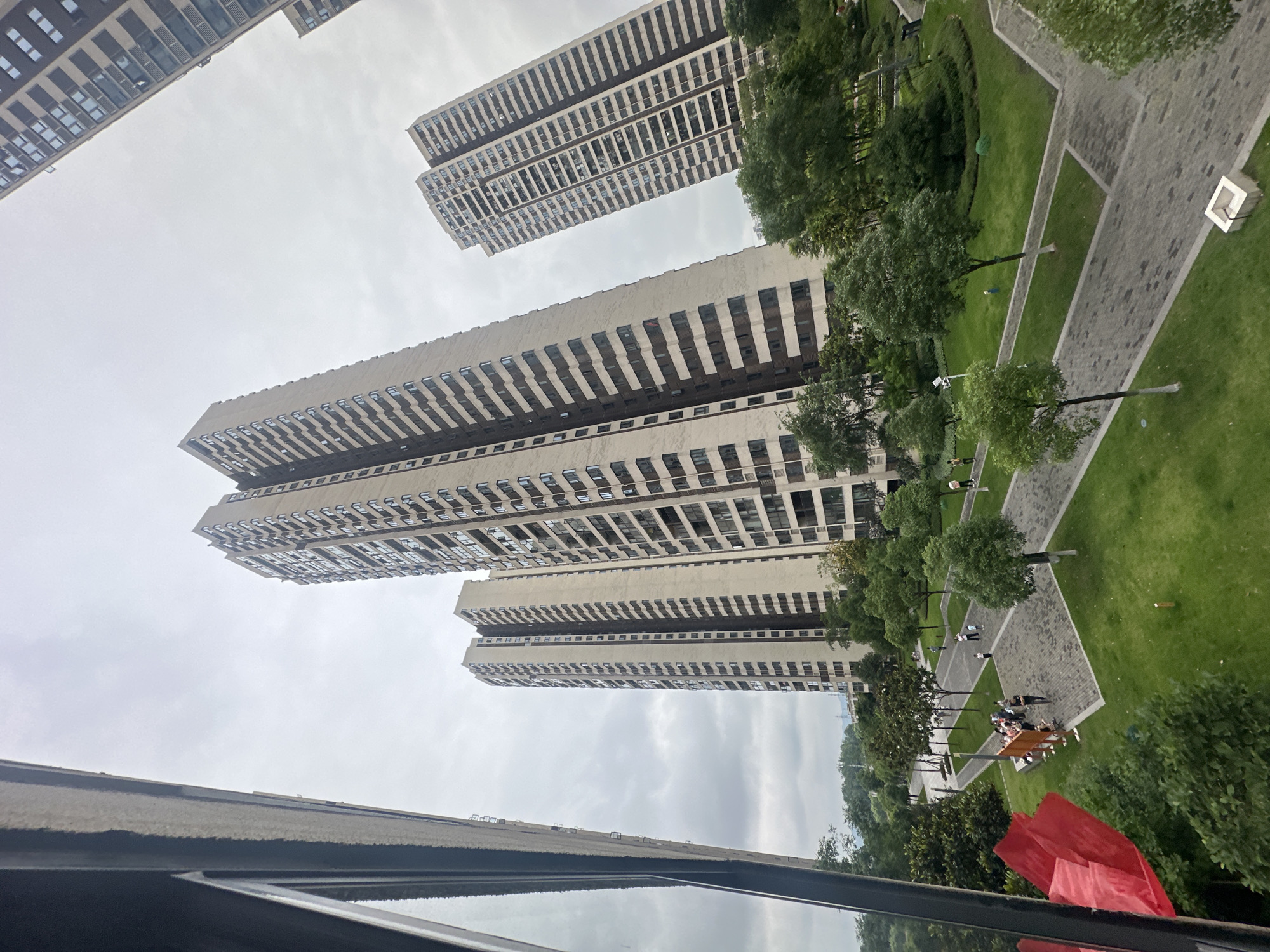 The image shows a high-angle view of a cityscape in Chongqing High-tech Zone, China, with several tall, residential apartment buildings dominating the scene. The buildings are uniformly designed with multiple rows of windows, and some have dark horizontal accents. A green space is visible, with trees, shrubs, and grassy areas interspersed with gray paved walkways. Groups of people are present along the walkways. An overcast sky and reflective surface (window) are in the foreground.
