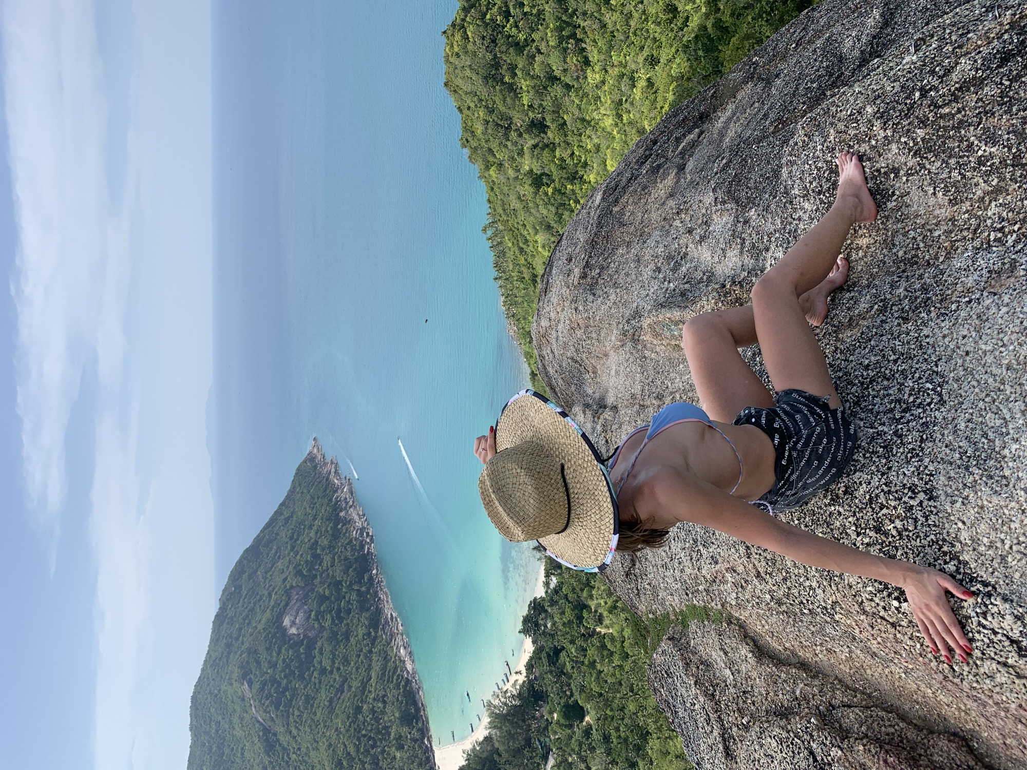 A young woman wearing a straw hat and a blue bikini top with black shorts sits on a rocky outcrop, looking out at a scenic bay. The bay features a crescent-shaped white sand beach, turquoise water, and lush green jungle-covered hills. Several small boats are visible in the shallow water, and a boat is leaving a white wake further out in the ocean. The sky is clear with a few wispy clouds. The scene appears to be taken from a high vantage point, likely a viewpoint or overlook in the Ko Pha-ngan District of Thailand.