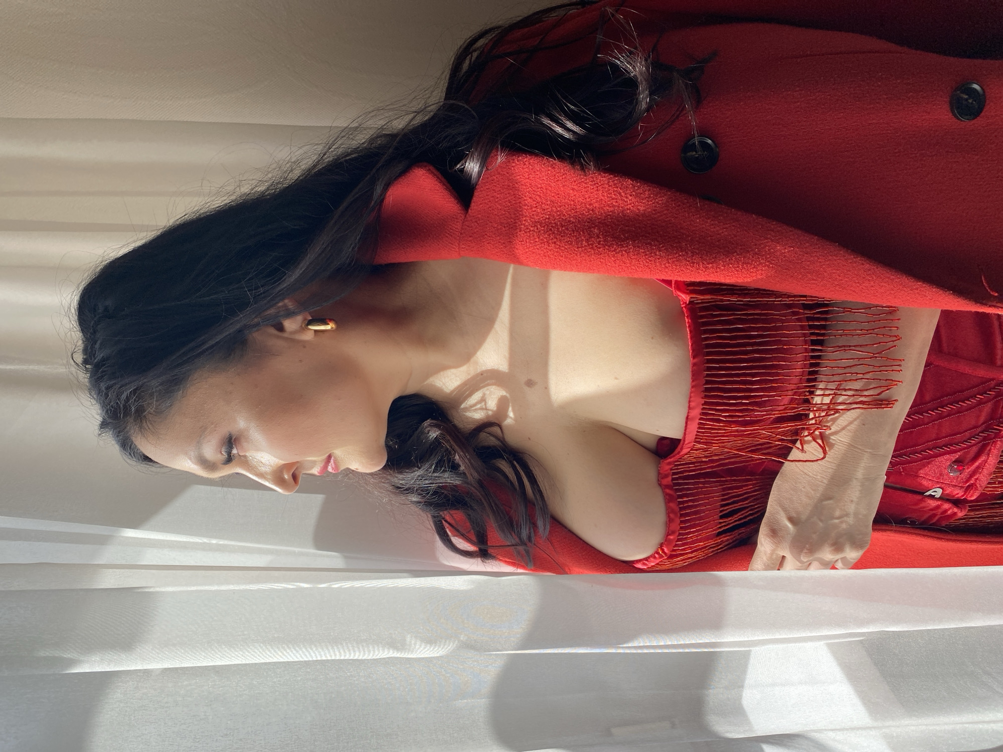 A medium shot depicts a woman facing left, with her head positioned towards the bottom left of the frame. She wears a red garment with fringe and a red coat. The background consists of white fabric. The location is not discernible from the provided image, and therefore the city of Yekaterinburg, Russia, cannot be confirmed.