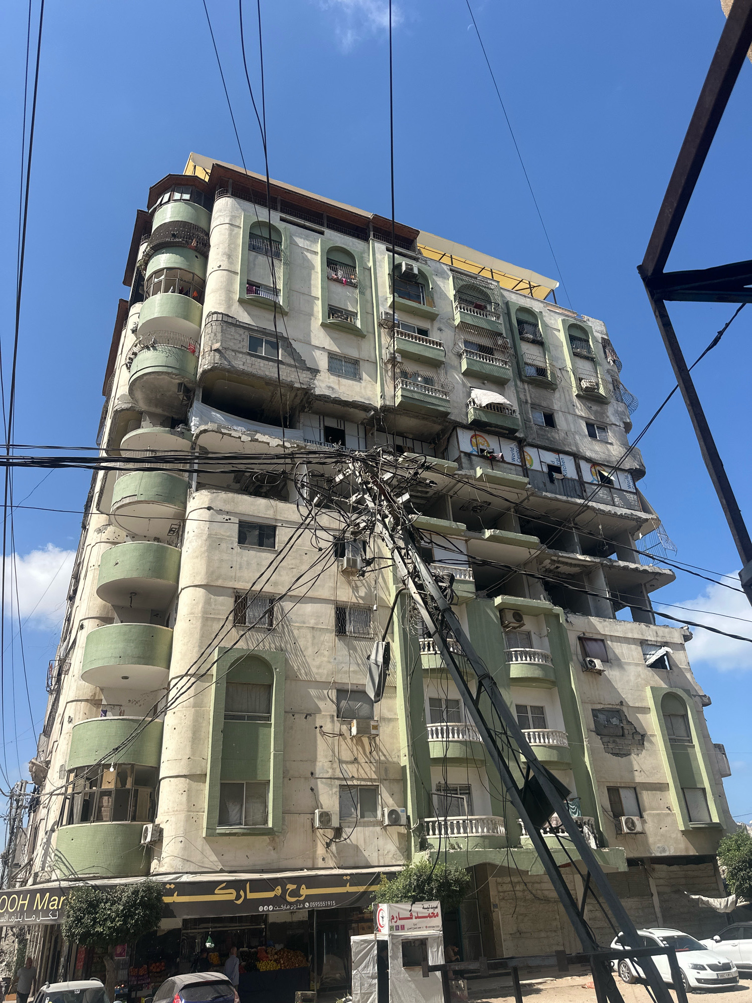 An exterior, low-angle, eye-level shot captures a partially damaged, multi-story apartment building in the city of Gaza, Palestinian Territories. The building, painted in pale green and cream, exhibits evidence of structural compromise, including missing sections of walls, exposed rebar, and debris scattered across balconies and ledges. Numerous windows are visible, some with curtains, others broken or boarded up. Air conditioning units are mounted on the exterior walls.
A complex network of overhead electrical wires crisscrosses the foreground and drapes over the building, with one utility pole leaning precariously. The sky above is a clear, bright blue with a few wispy clouds.
At the ground level, a storefront with a dark awning displays signs in Arabic. The signs include "OOH Market" in English and what appears to be "سوق ماركت" in Arabic script, along with phone numbers. In front of the market, a display of fruits and vegetables is visible, and several people are standing outside or near vehicles parked on the street. A small white tent with red lettering and a crescent symbol is also visible near the storefront. The overall impression is one of urban life in an area experiencing significant damage.