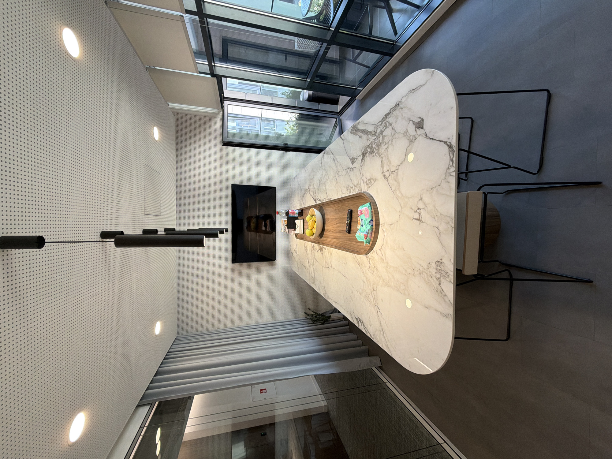 In an indoor setting, a large, elongated marble conference table with a polished finish dominates the center of the frame. The table has rounded edges and a distinctive, organic shape, supported by thin, black metal legs. On the table, a wooden tray holds a bowl of yellow fruit, a bottle of soda, and a stack of packaged wet wipes. A remote control rests near the wipes. Behind the table, a wall-mounted television displays an image of a food spread. Suspended from the white, perforated ceiling are three cylindrical black pendant lights. To the left of the table, a pale blue curtain is drawn, partially obscuring a glass-partitioned room. On the right, large windows with dark frames look out onto a balcony with chairs and a table. The floor is a dark gray, matte material. The overall impression is that of a modern, high-end meeting or dining space.