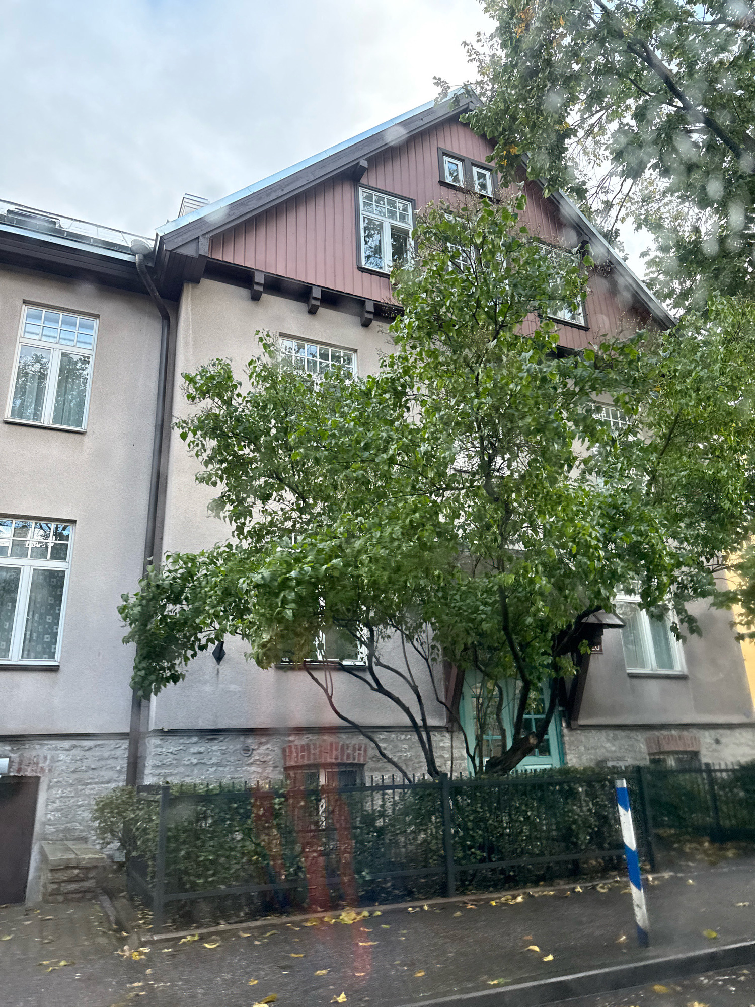 The image shows a street view of a building in Tallinn, Estonia, on a cloudy day. The building is a multi-story structure with a light-colored facade and a reddish-brown upper story with wooden siding. There are several windows with white frames visible on the building. A large tree with green leaves partially obscures the building. In front of the building, there is a black metal fence with a hedge behind it. The ground in front of the fence is a paved area with fallen yellow leaves scattered on it, suggesting it is autumn. A blue and white striped pole is visible on the right side of the image, near the edge of the paved area. The overall impression is that of a residential street scene in a European city during the fall season.