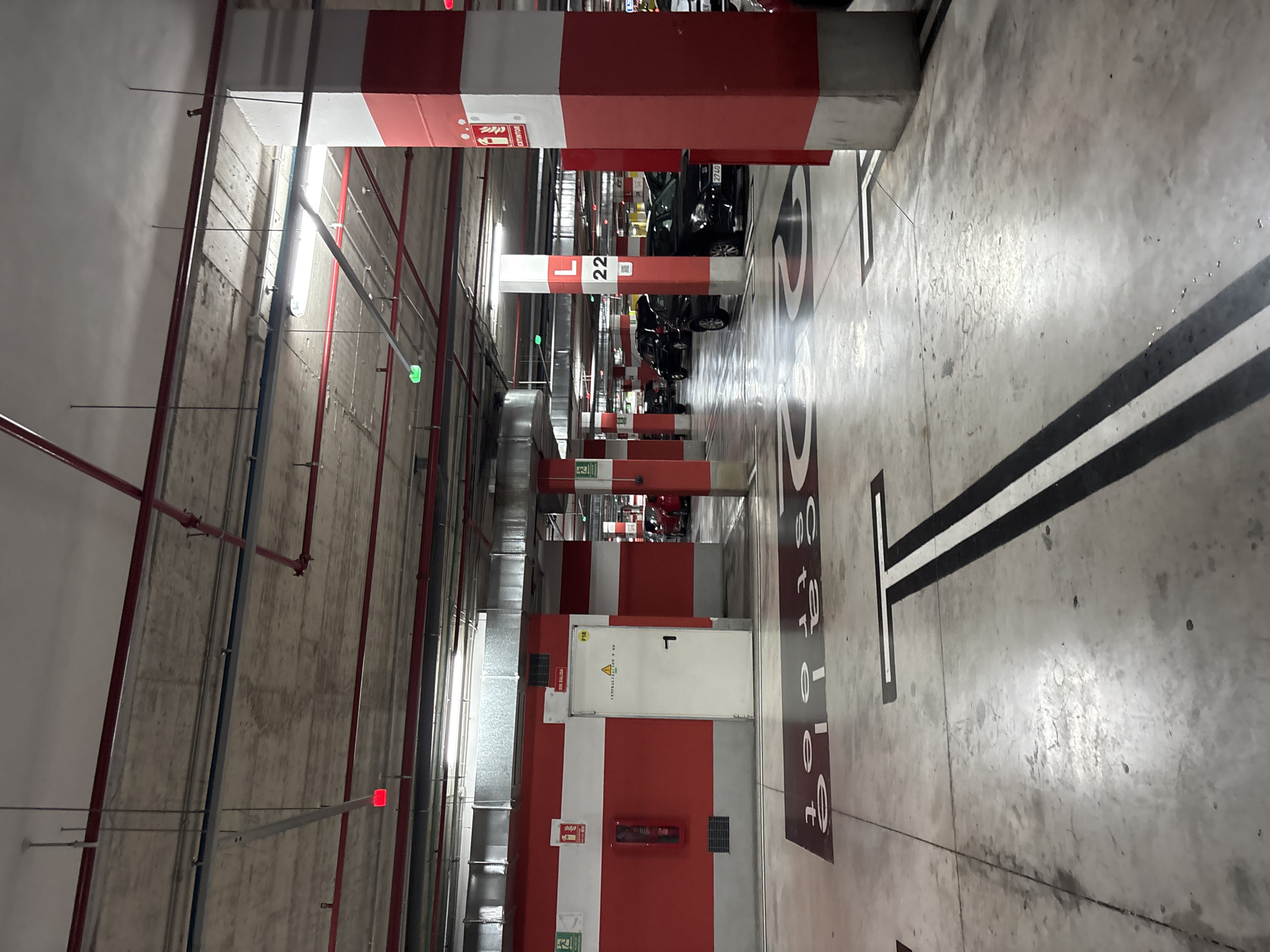 The image depicts a brightly lit interior of a multi-story parking garage. The floor is concrete with painted markings indicating parking spaces and directional arrows. Large, red and white striped concrete pillars are spaced throughout the garage, supporting the concrete ceiling. The ceiling features exposed conduits, pipes, and fluorescent lighting fixtures. Red emergency lights are visible on the ceiling and on the walls. On the left side of the image, a white door is set into a red and gray wall, with a fire extinguisher mounted to the left of the door. The words "SIN SALIDA" (No Exit) are written above the door. The number "F18" is also visible above the door. Further down the aisle, cars are parked in designated spots. A sign on one of the pillars indicates "L 22" and has a QR code. The overall environment suggests a functional, well-maintained urban parking facility. Given the architectural style and signage, this could be located in the city of Orihuela, Spain, a region known for modern infrastructure. There are no people clearly visible in the image.