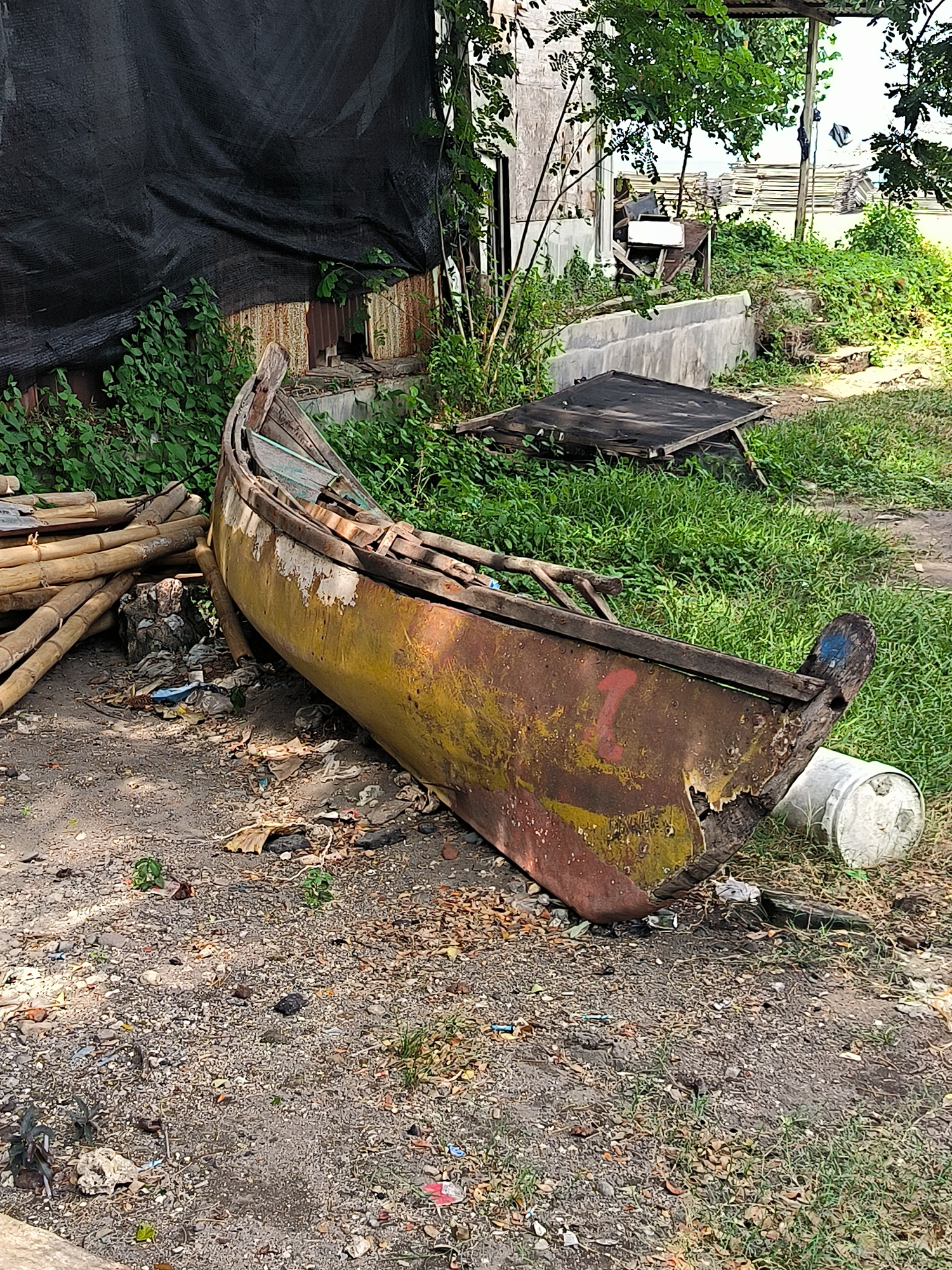 The image depicts a disused wooden boat, heavily weathered and rusted, lying on a patch of dirt and overgrown grass. The boat is positioned diagonally, with its stern facing towards the right side of the frame. Its exterior is a mix of peeling yellow paint and exposed rusty metal, indicating significant age and neglect. To the left of the boat, a pile of bamboo poles is stacked haphazardly, partially obscuring some scattered debris and trash on the ground.
In the background, a weathered building with corrugated metal siding is visible, partially covered by a large black tarp. Lush green vegetation, including trees and shrubs, grows around the building and the boat, suggesting an outdoor, possibly semi-abandoned, location. A white bucket is placed near the stern of the boat. The ground itself is a mixture of dirt, small stones, and scattered leaves, with more trash visible in places. The overall impression is one of disrepair and abandonment.
This scene is located in the city of Lhokseumawe, Indonesia. The environment suggests an area that might be near a coastline or a waterway, given the presence of a boat, though it is currently far from any visible water. The combination of old structures, natural overgrowth, and discarded materials points to a possibly industrial or semi-rural fringe area of the city. There are no people or animals visible in the frame.