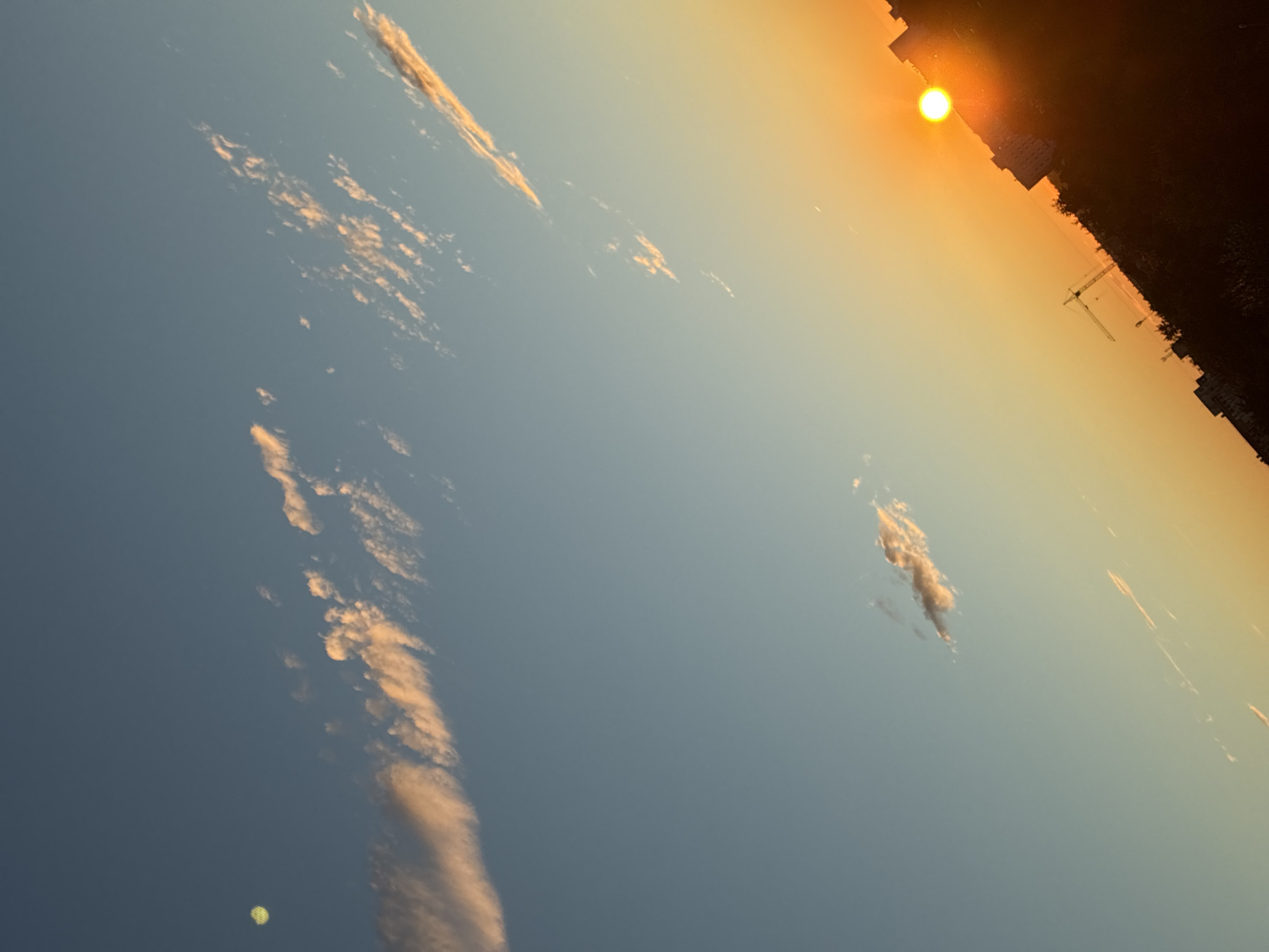 The image shows a sky with clouds illuminated by the setting sun. The sun, a bright orange orb, is low in the sky, casting a warm glow. Silhouettes of buildings and trees are visible against the vibrant sky, suggesting an urban environment. A construction crane can be seen in the distance. The image is taken from a high vantage point, looking down on the city. The sky transitions from a clear blue at the top to a warm orange and yellow near the horizon. Wispy clouds are scattered across the sky, catching the sunlight.