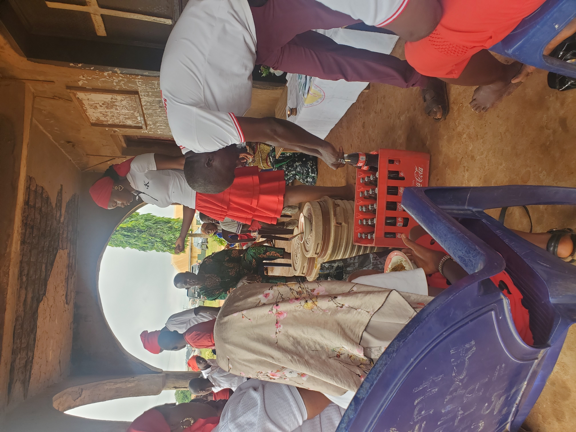 This is a medium-shot, eye-level, outdoor photograph taken in Nsugbe, Nigeria, at what appears to be a communal gathering or event, possibly religious. The scene is set under an open-air structure with concrete archways and a rough-textured ceiling. The ground is a dirt or sandy surface.
In the foreground, a person wearing a red top and dark sandals is seated on a blue plastic chair, partially visible. To the right, another individual in a white polo shirt with red trim and dark maroon pants is bending down, reaching for a Coca-Cola bottle from a red crate. Their left hand is holding the neck of a bottle.
Behind them, a woman in a white polo shirt with a black logo and a tiered red skirt is standing. She has a red headscarf and wears large hoop earrings. To her left, a person in a green patterned outfit is seated, appearing to be an elder, looking towards the gathering. Various other individuals, including children, are visible in the background, some wearing red head coverings.
Several objects are present, including a crate of Coca-Cola bottles, stacks of round, covered containers (possibly for food), a white tablecloth draped on a table with some papers or brochures, and a small lit candle. A wooden cross is affixed to the wall in the background. The overall atmosphere suggests a casual, community-oriented event.