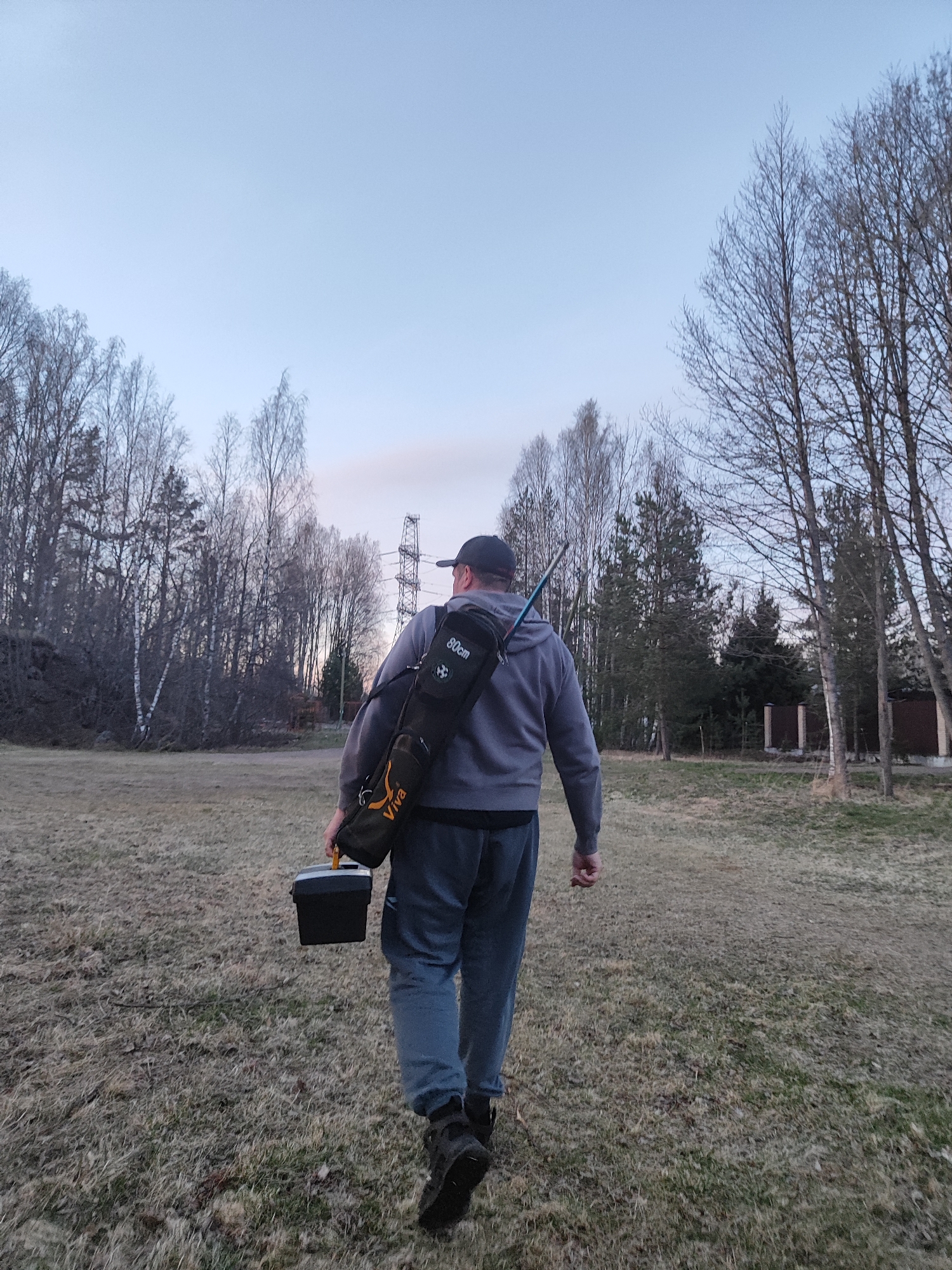 A person is shown from behind, walking across a field of dry, sparse grass. They are wearing a dark baseball cap, a grey hooded sweatshirt, dark sweatpants, and dark boots. On their back, they carry a black and orange shoulder bag, which appears to be an equipment case, possibly for fishing rods, with a long blue object protruding from the top. Visible text on the bag includes "80cm" and "Viva" with a small emblem. In their right hand, they hold a small, black rectangular container with an orange handle, resembling a tackle box or cooler.
The scene depicts an outdoor, natural environment, likely a rural or semi-rural area. A dense treeline composed of bare deciduous trees and evergreen conifers forms the background. In the distance, a large metal power line tower stands among the trees. On the far right, a glimpse of a building or house structure with a fence is visible.
The sky is a clear, light blue, suggesting either late afternoon or early morning light. The bare branches of the deciduous trees indicate either late winter or early spring. The activity appears to be an individual embarking on an outdoor pursuit, likely fishing or a similar recreational activity, given the equipment. The overall mood is quiet and calm.