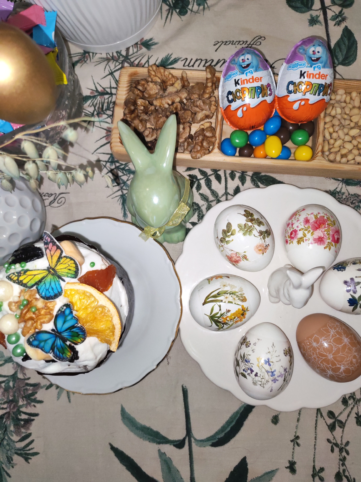 The image shows a tabletop Easter display. A decorated Easter cake with butterfly embellishments and orange slices sits on a white plate. Next to it, a white plate holds several decorated Easter eggs and a small white bunny figurine. A wooden tray contains walnuts, chocolate candies, Kinder Surprise eggs, and peanuts. A green ceramic bunny figurine stands nearby. Other items, including a golden egg and floral arrangements, are also present. The setting seems to be indoors, possibly a home in Izhevsk, Russia.