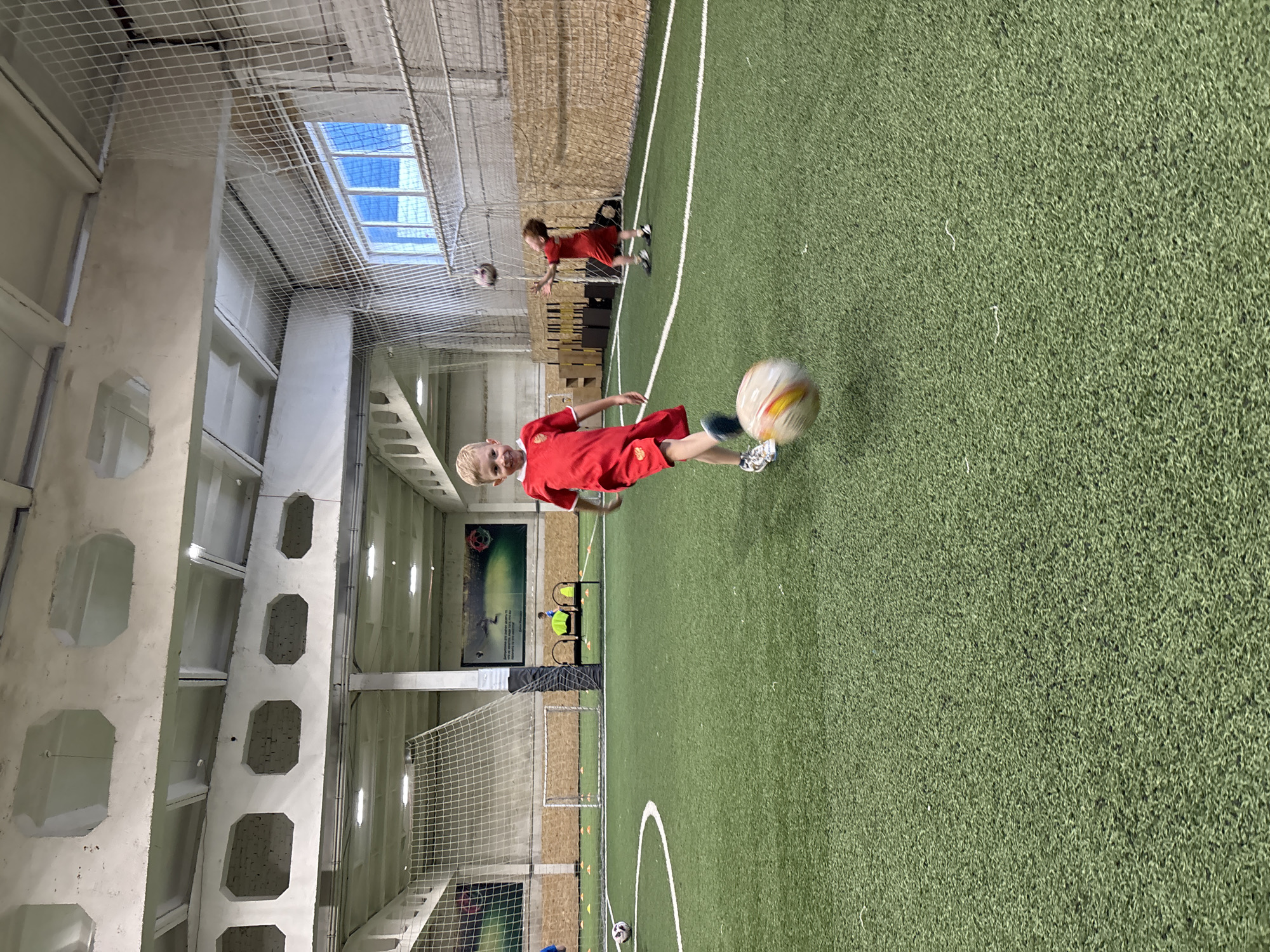 A young boy in a red soccer uniform kicks a soccer ball on an indoor artificial turf field. He is in the foreground, facing the camera with a slight smile. The ball is in mid-air, blurred from motion. In the background, another child in a similar uniform is tossing a soccer ball in the air. The field has white markings and orange cones are visible near a soccer net. The environment appears to be a sports facility, possibly a children's soccer academy, located in Chaikovsky, Russia, evidenced by the architectural style and typical indoor sports field construction. The ceiling is high, with exposed concrete beams and fluorescent lighting. A large mural depicting a soccer-related image is on the far wall.