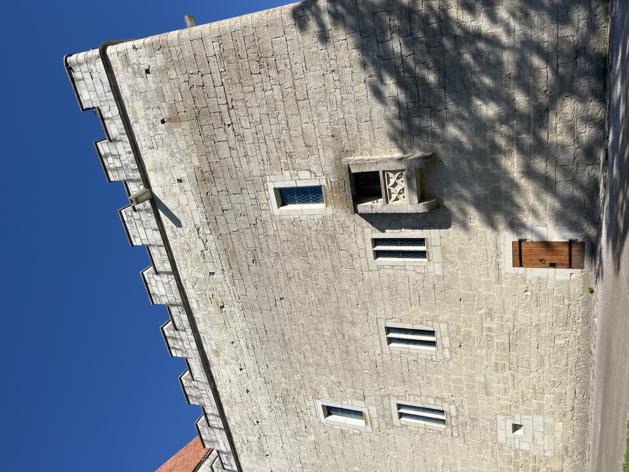 An exterior, low-angle, wide shot shows the stone wall of Kuressaare Castle in Kuressaare, Estonia, under a clear blue sky. The wall is constructed of roughly hewn, light-colored stone blocks and features a crenellated parapet along the top. Several narrow, rectangular windows with diamond-patterned glass panes are visible. To the right of the center, a small balcony juts out from the wall, supported by decorative corbels. A wooden door with iron hardware is set into the wall near the bottom right. Shadows of tree branches are cast across the lower portion of the wall. The ground in front of the castle is a gravel path.