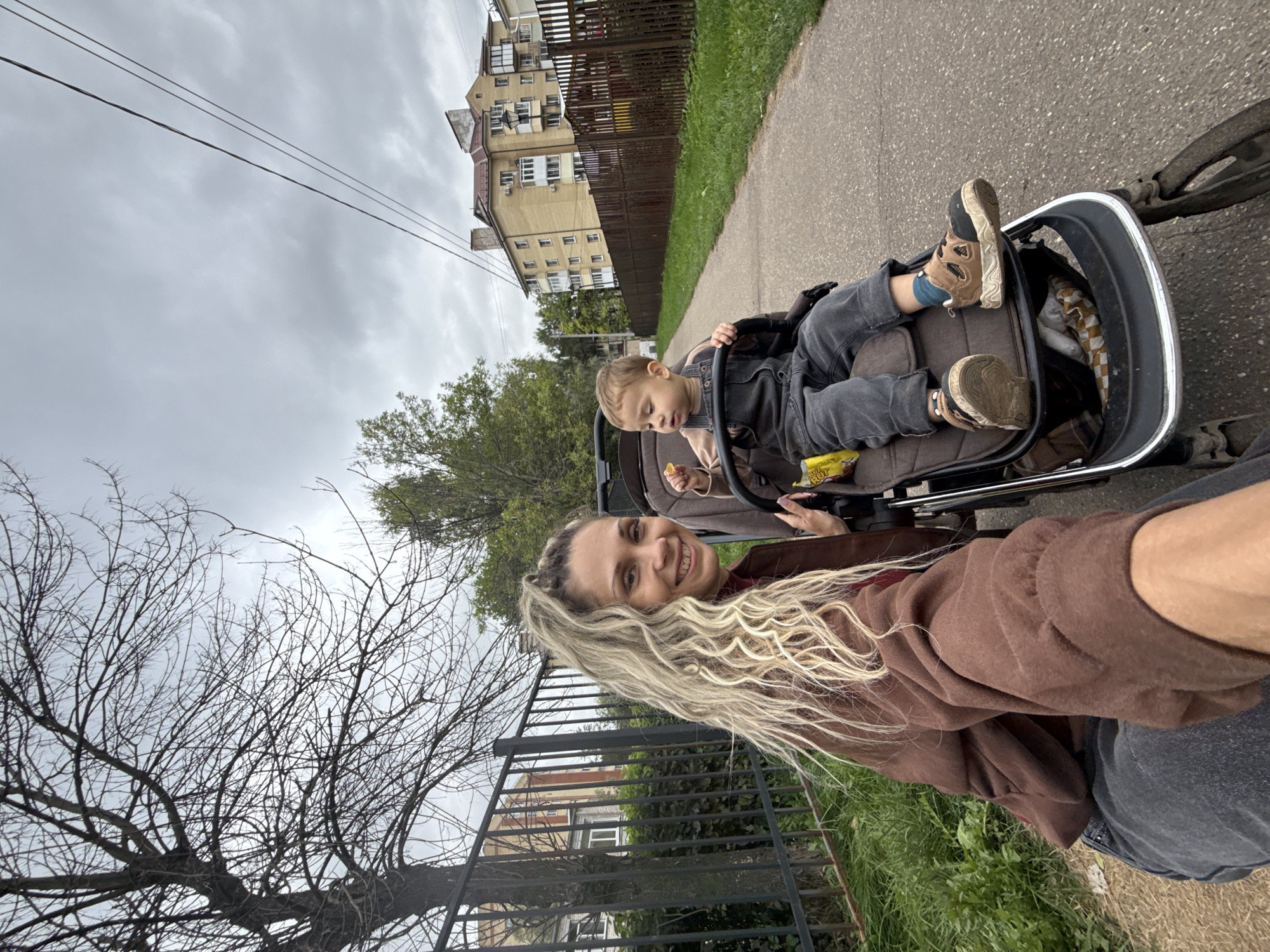The image depicts a woman and a young child in a stroller on a paved path in a residential area. The woman, with long, curly blonde hair and a brown jacket, is smiling and holding the stroller. The child, wearing a denim jumpsuit, is seated in the stroller and holding a yellow object. In the background, there are apartment buildings, a fence, and trees under a cloudy sky. The overall setting suggests an outdoor urban environment in Rusanovo, Russia.