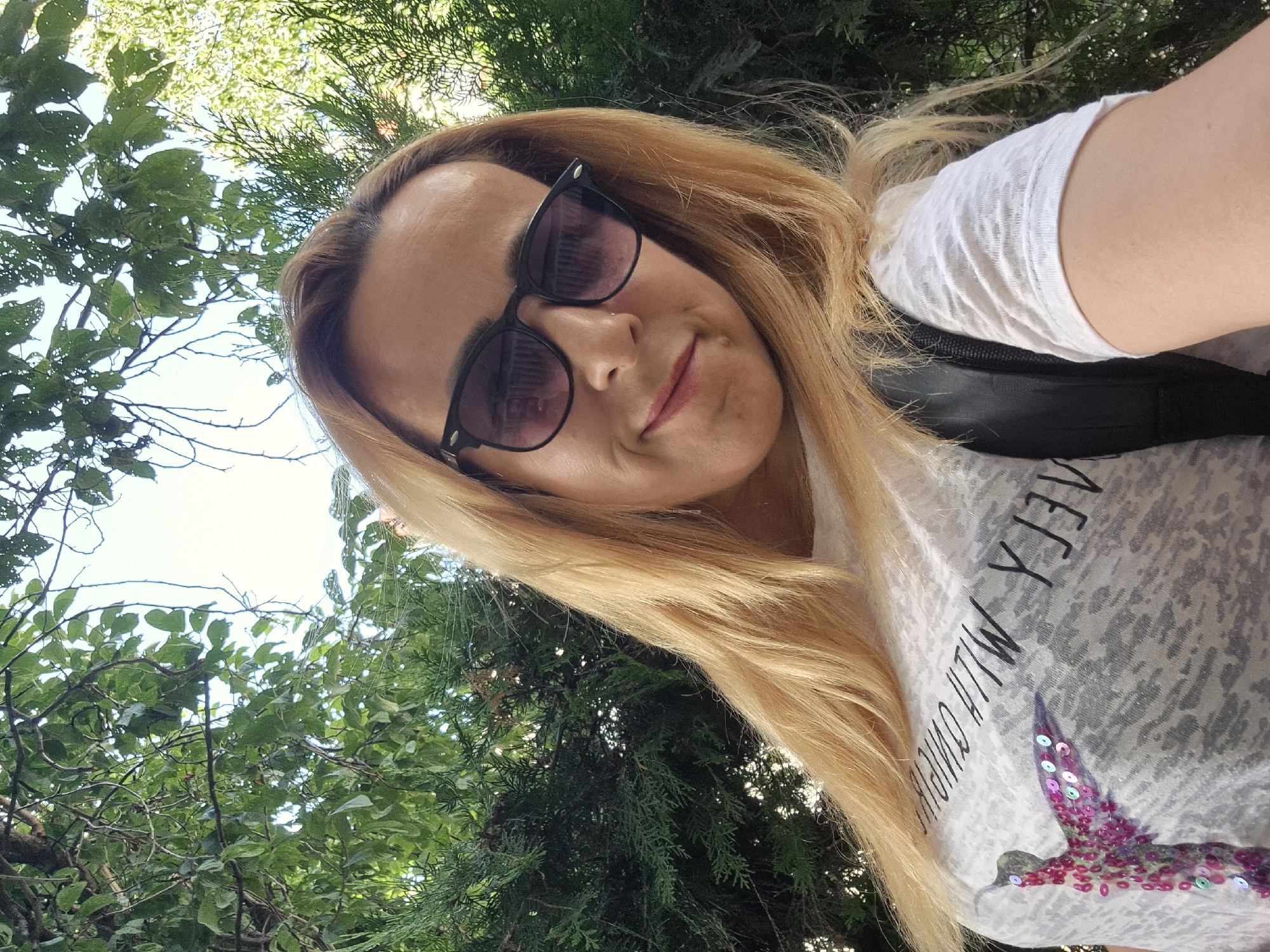 A woman with long blonde hair and sunglasses takes a selfie outdoors, surrounded by lush green trees. She is wearing a white t-shirt with a design featuring a bird decorated with pink and purple sequins. Visible text on the t-shirt appears to read "FLY WITH CHICKS". The lighting suggests it is daytime, with sunlight filtering through the leaves. The woman has a slight smile, giving a casual and relaxed impression. The camera angle is upward, typical of a selfie taken outdoors.