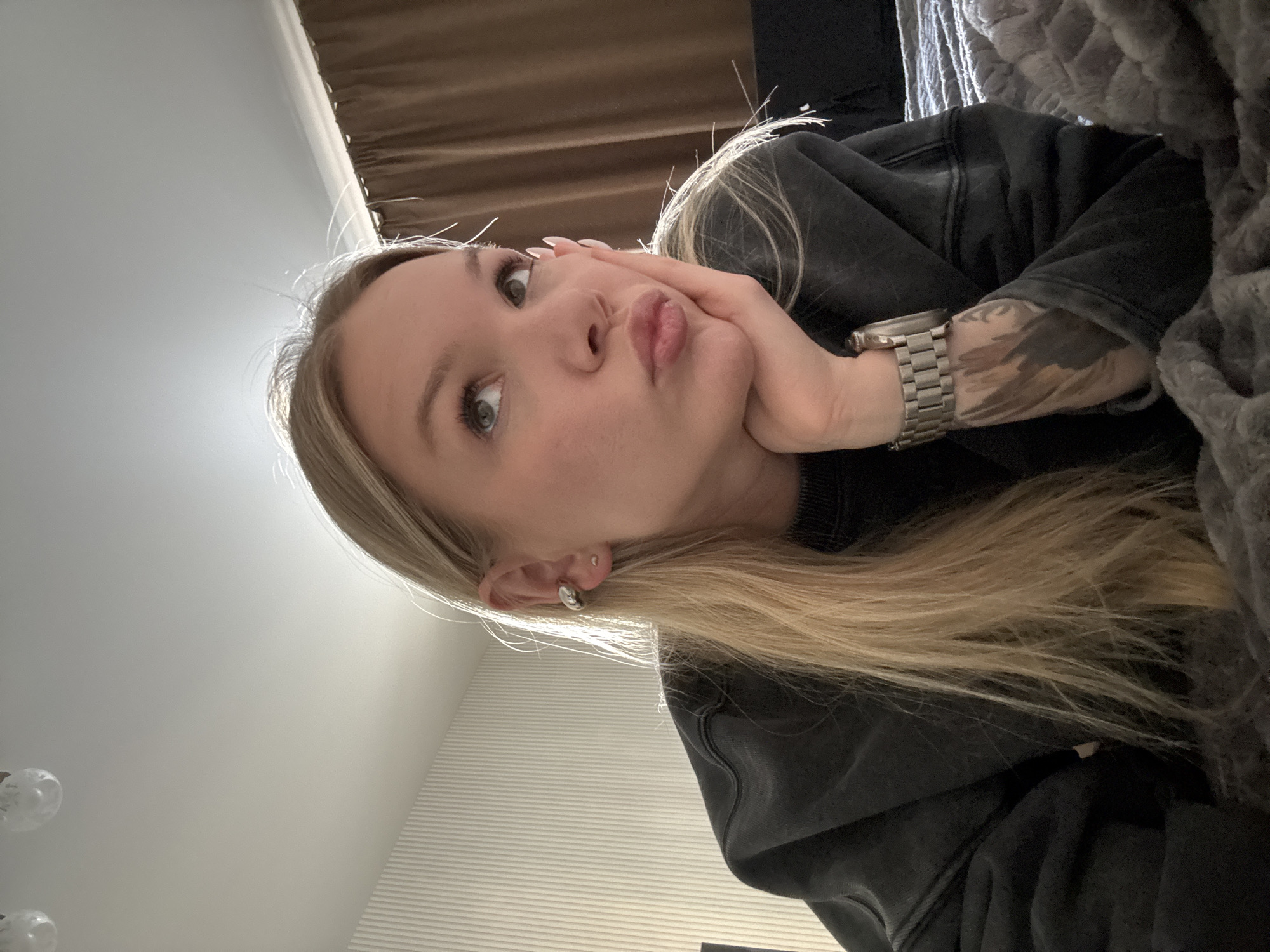 A young blonde woman is lying on a gray blanket. She is wearing a black hooded sweatshirt and a silver watch on her left wrist, which has a tattoo on it. Her right hand is supporting her face, and she is pursing her lips. The background includes a brown curtain and a white wall. The location is Perm, Russia, though this cannot be confirmed from the image alone.