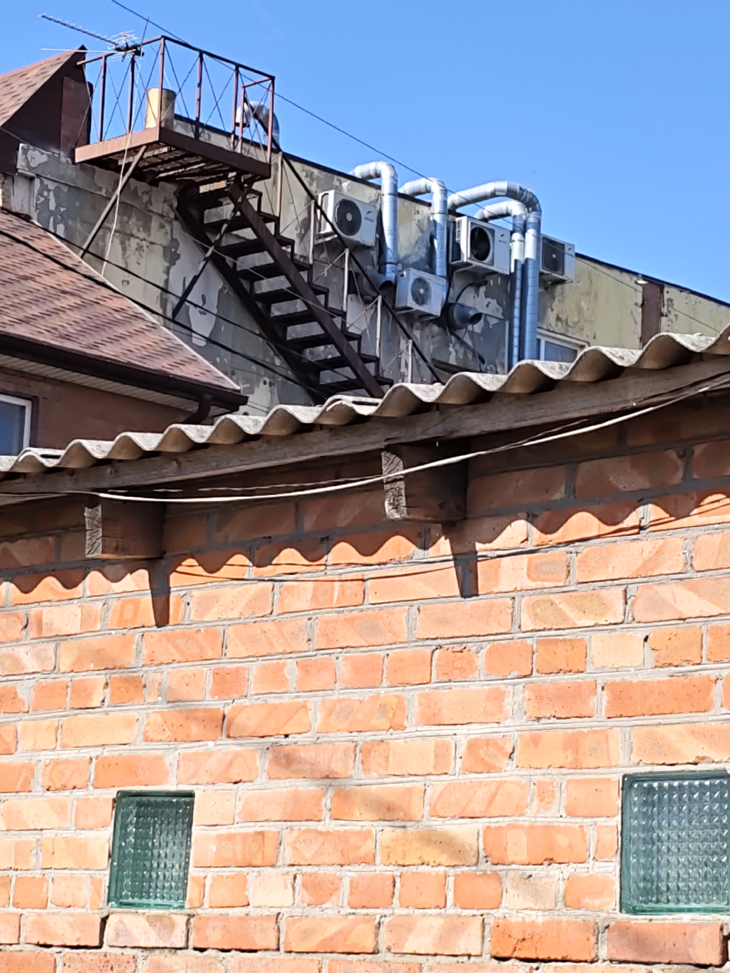 This is a medium shot, exterior photograph of the upper section of a brick building in Rostov, Russia, under a clear blue sky. The building features a red-tiled roof on the left, with a dark brown edge. Adjacent to the roof is a weathered, light-colored wall with peeling paint. A metal fire escape with a landing and stairs is attached to this wall. On the roof of the main building, several outdoor air conditioning units are visible, connected by silver pipes. A corrugated asbestos roof extends from the right side of the frame, supported by wooden beams, casting shadows on the brick wall below. The brick wall itself is made of orange-red bricks with gray mortar. Two small, frosted glass windows are set into the brick wall. The scene appears to be a utilitarian part of a building, possibly an industrial or commercial structure, with no people visible.
