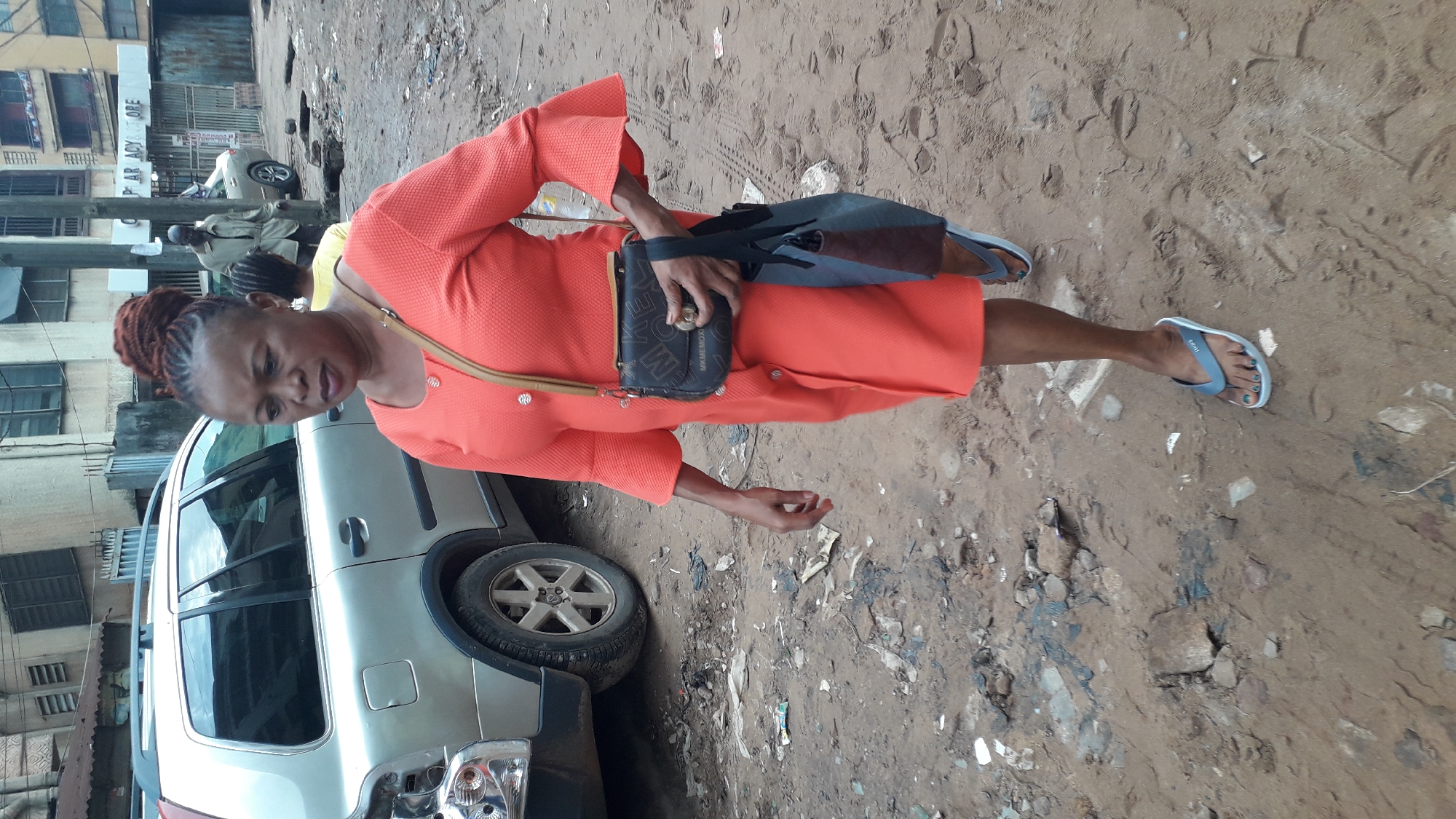 A woman wearing an orange dress and blue flip-flops walks on a dirt and debris-covered ground next to a silver SUV. She carries a dark purse with a strap over her shoulder and holds a black bag in her hand. The location appears to be a city street in Onitsha, Nigeria, as the background includes buildings and structures typical of urban environments in the region, with possible shops and residential units visible. The scene suggests a mundane moment in the city, possibly related to shopping or transportation.
