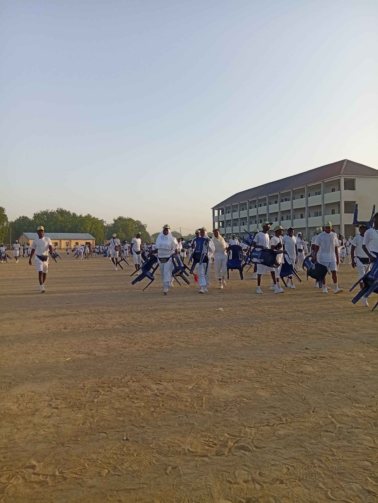 The image shows a group of people in white clothing and green caps carrying blue chairs across a sandy field. There is a large building in the background, and trees can be seen in the distance. The ground is covered with footprints. The location is in Maiduguri, Nigeria.