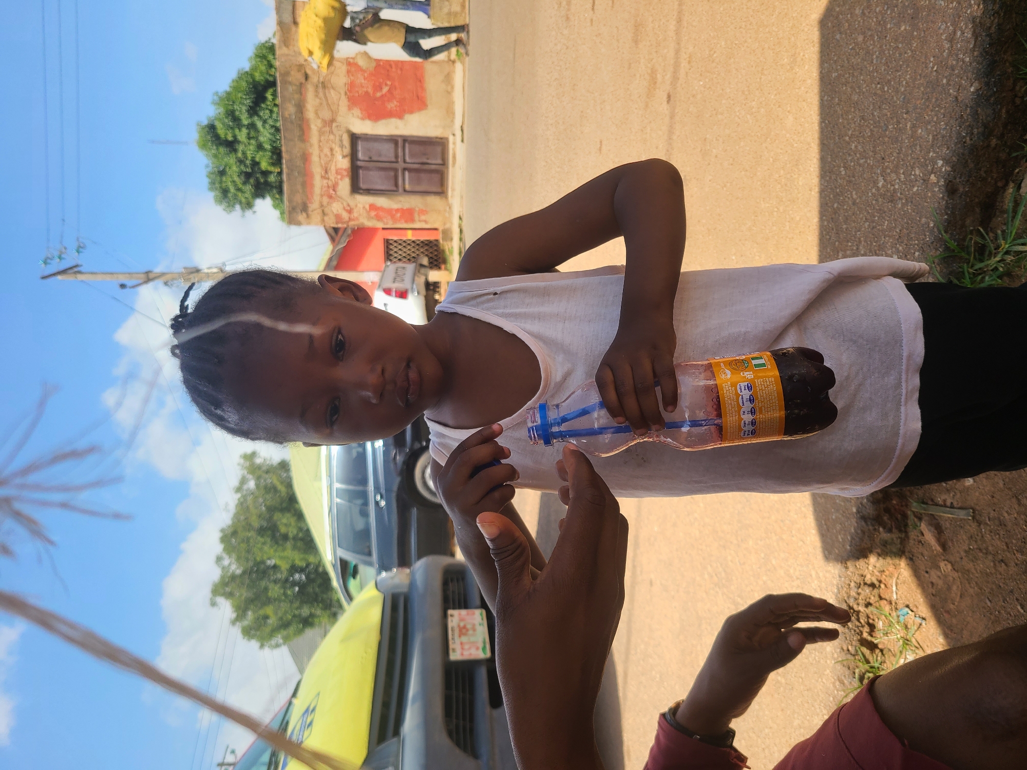 In the city of Jos, Nigeria, under a clear sky with scattered clouds, a young girl in a white tank top and black pants stands holding a bottle of dark-colored liquid with a blue straw. Her braided hair has a white streak. An adult's hand, partially visible on the left wearing a red garment, is interacting with the girl, appearing to offer or receive something. In the background, a yellow vehicle and a dark SUV are parked on a street. Further back, a building with red and brown stucco walls and wooden double doors is visible, with two people carrying yellow objects on their heads walking past. The ground is paved with a sandy-colored surface, and sparse green grass grows along the edge of the street.
