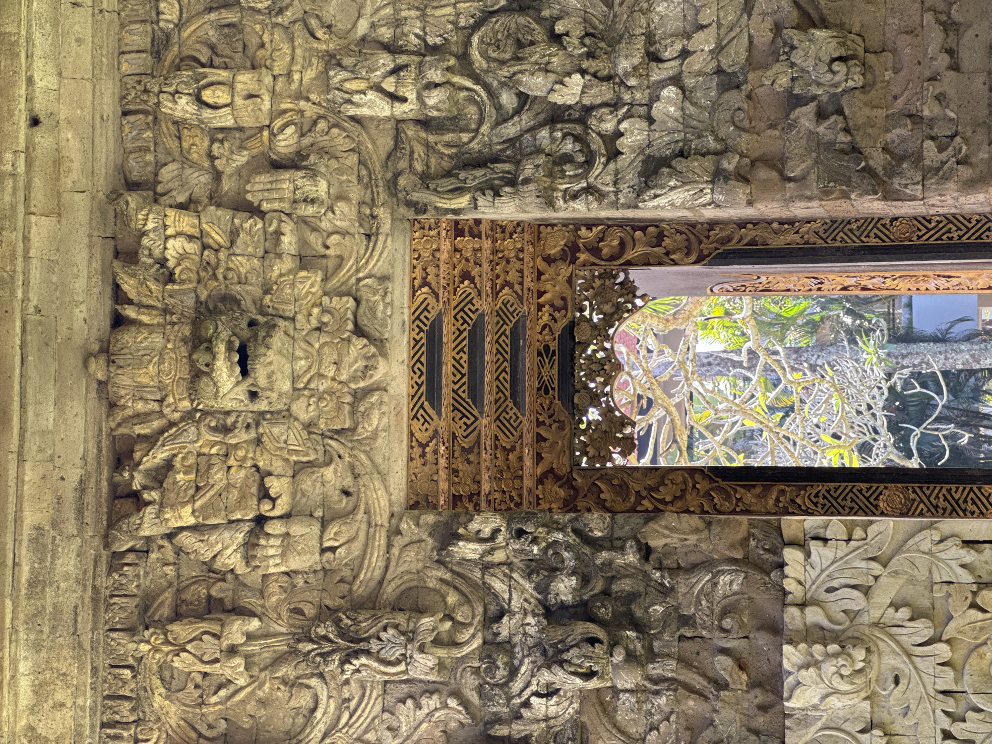 The image depicts a stone wall with intricate carvings and a window in Kuta Selatan, Indonesia. The wall is made of light-colored stone with detailed relief carvings of floral and animal motifs. The window has a wooden frame with decorative carvings, and through it, lush green vegetation is visible. The scene is well-lit, highlighting the textures and details of the stonework and woodwork.