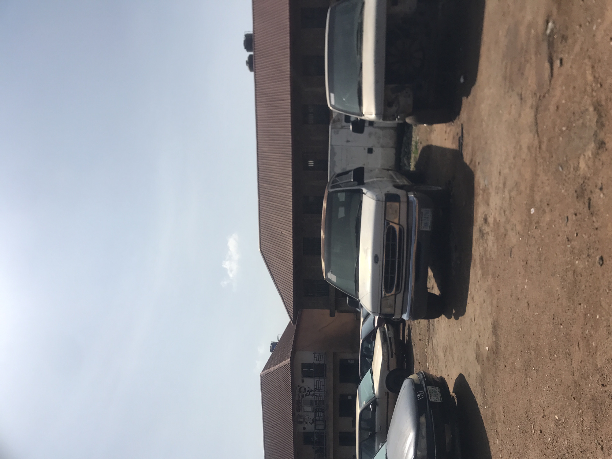 The image captures an outdoor scene in what appears to be a car repair or storage lot in Rafin Par, Nigeria. In the foreground, several vehicles are parked on a dirt and gravel surface. A white van, possibly a Ford E-Series, is prominently featured in the center, with its front grille and headlights visible. To its right, another white vehicle, also appearing to be a van, is partially visible and seems to be missing its front bumper and headlights. To the left of the central van, a dark-colored sedan is parked, with its front bumper and license plate clearly discernible, displaying the text "BS4-43CM". Further in the background, several other cars, including a light-colored sedan, are parked.
Behind the vehicles, two multi-story buildings with reddish-brown corrugated metal roofs dominate the scene. The building on the left has a light-colored facade with balconies and windows. The building on the right has a similar roofline and appears to be constructed from darker materials. The sky is mostly clear with some light cloud cover, suggesting daytime. The overall environment appears to be somewhat unkempt, consistent with a working lot. No people are visible in the image.