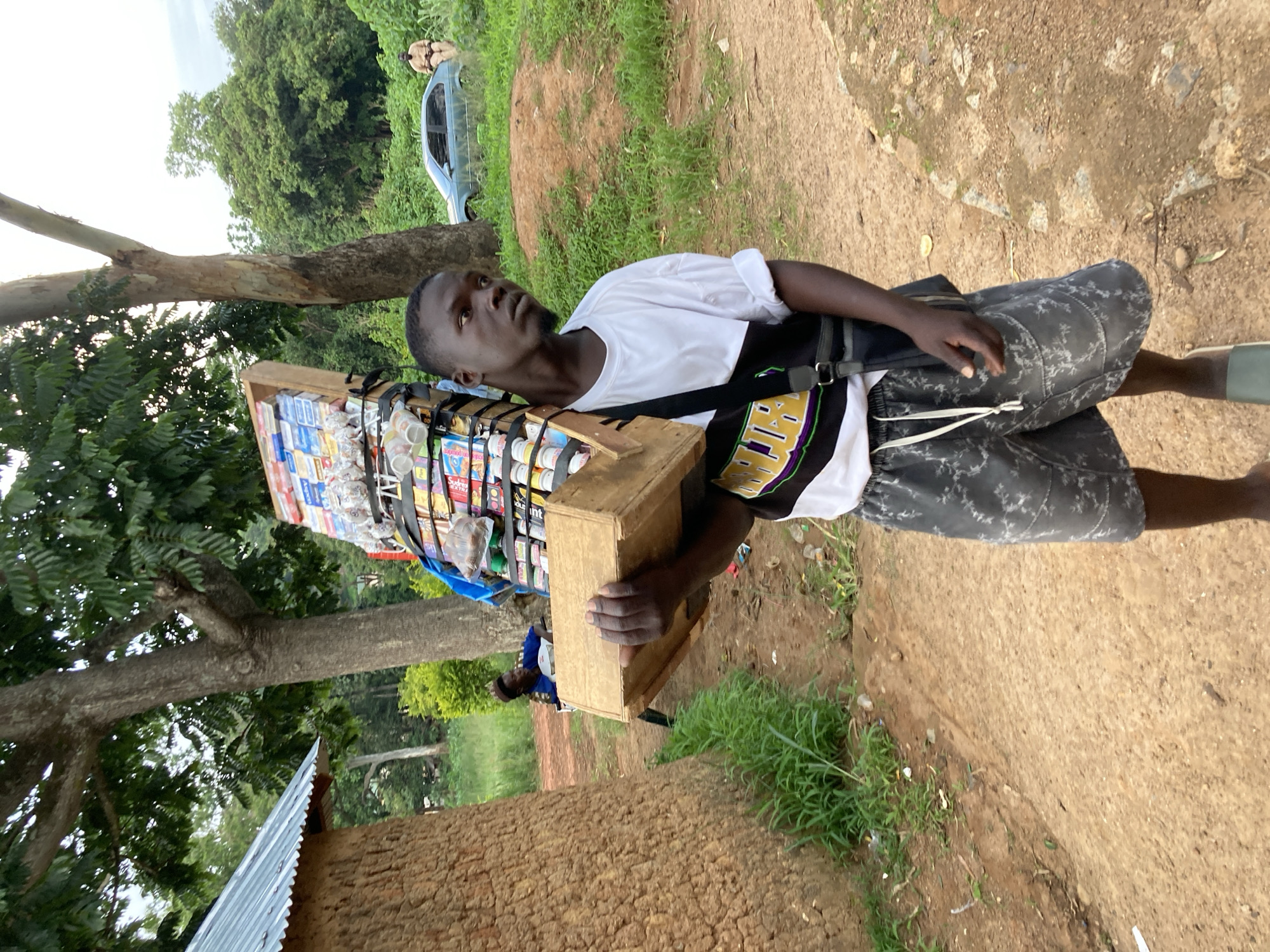 A young Black man, likely in his late teens or early twenties, is walking outdoors in a dusty, unpaved area, possibly on the outskirts of Jos, Nigeria. He is carrying a wooden crate filled with various packaged goods, including small bottles, cans, and boxed items, balanced on his left shoulder. He wears a white t-shirt with black and yellow accents on the chest, dark patterned shorts, and dark-colored slides. A black crossbody bag is slung over his right shoulder. The background features lush green vegetation, large trees, and a rudimentary mud building with a corrugated metal roof. In the distance, a light blue car is parked on a dirt path, and another person is visible standing near it. The overall impression is of a busy marketplace or a community thoroughfare in a rural or semi-urban setting.