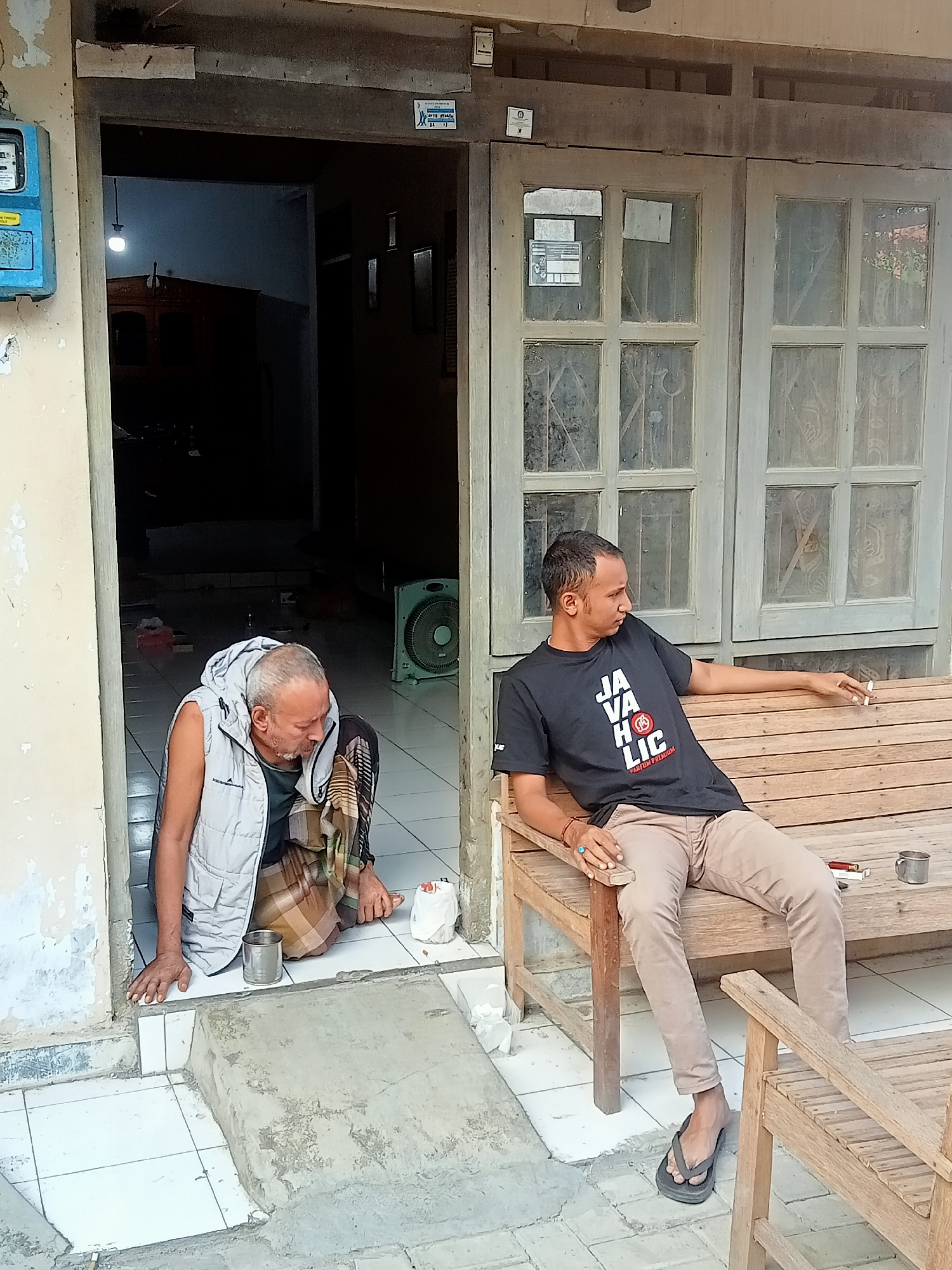 In Kecamatan Pati, Indonesia, two men are captured outside a building with a doorway and a multi-paned window. The man on the left, with graying hair and a beard, sits on the ground just inside the doorway, wearing a light gray vest over a darker shirt and a patterned sarong. He rests his left hand on the floor and his right hand near a metal cup. To his right, a younger man is seated on a wooden bench, smoking a cigarette and looking to the side. He is wearing a black t-shirt with white text that reads "JAVA HOLIC PREMIUM," beige pants, and dark flip-flops. A second wooden bench is visible in the foreground. A blue electrical meter is mounted on the wall to the left of the doorway. Inside the building, visible through the open doorway, a light bulb illuminates a dark interior with furniture and a floor fan. The outdoor area is paved with white tiles and concrete.